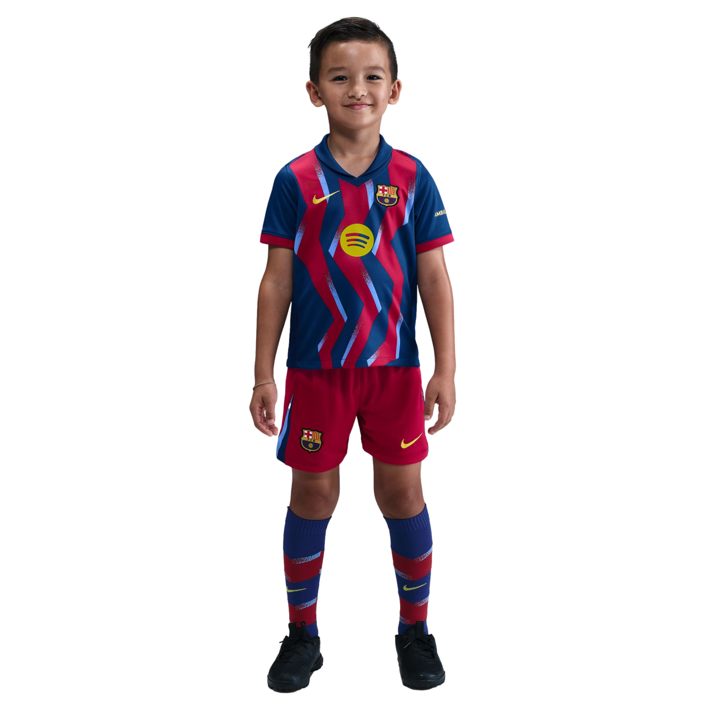 A young boy smiling in a blue and red zig-zag FC Barcelona soccer kit, including jersey, shorts, and socks, standing against a black background.