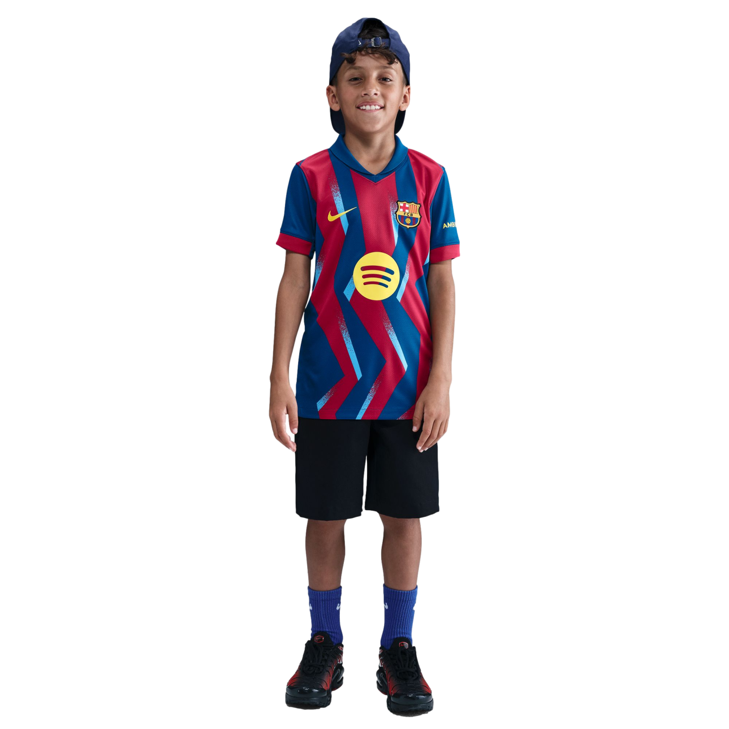 A smiling young boy wearing an FC Barcelona soccer jersey, black shorts, and a blue cap, standing against a solid black background.