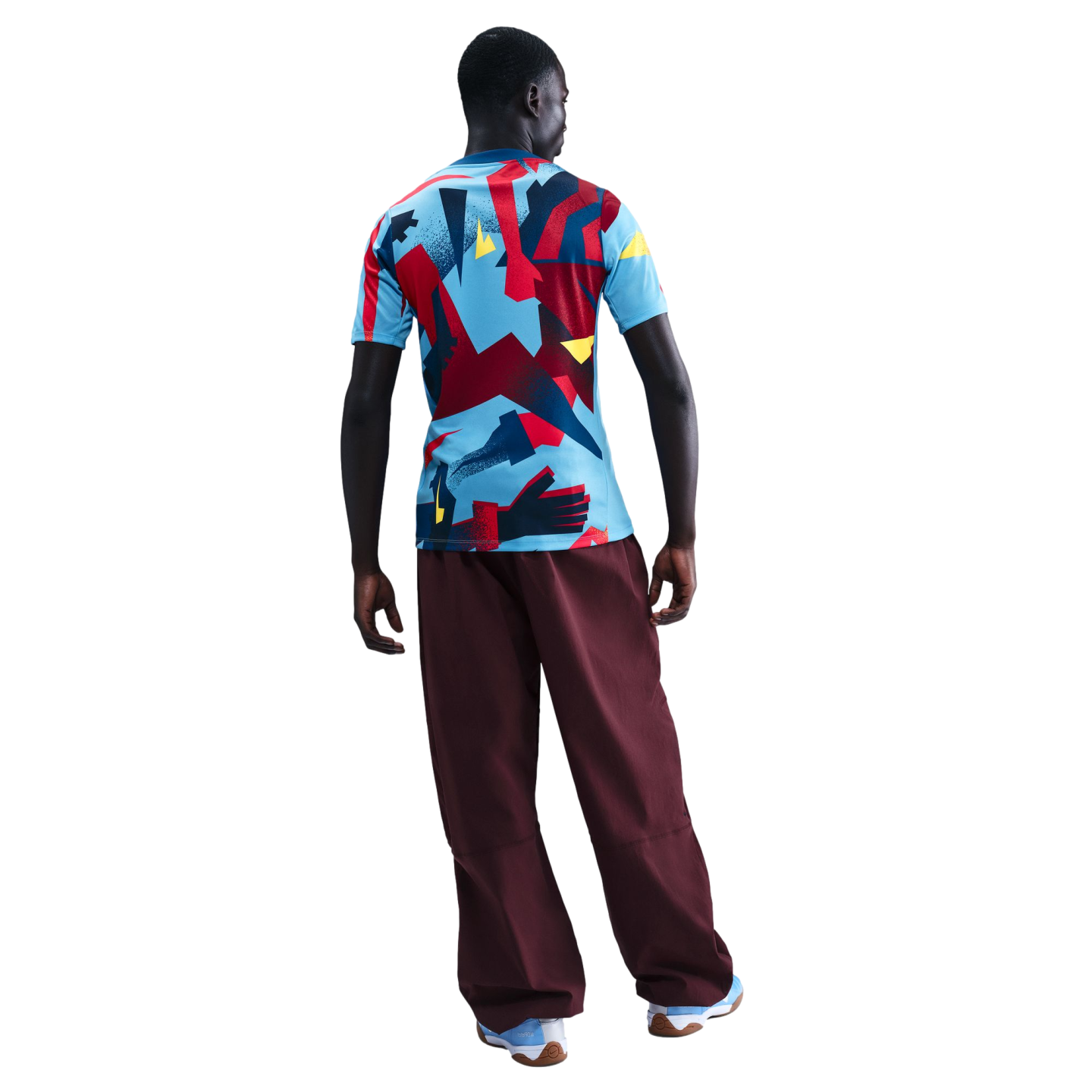 Back view of a person wearing a colorful geometric print T-shirt, loose maroon trousers, and light blue sneakers, standing against a black background.