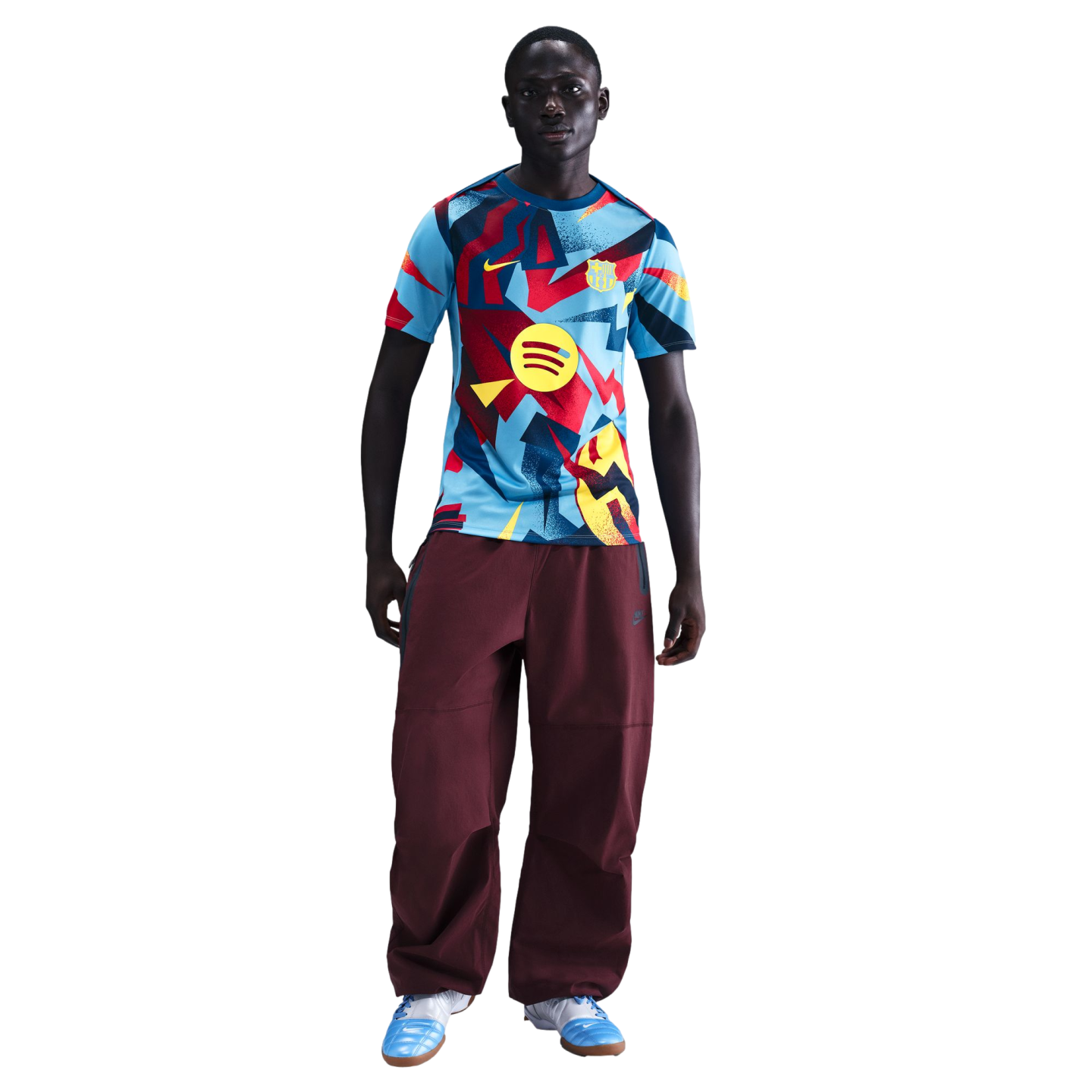 Man wearing a patterned FC Barcelona jersey, maroon baggy pants, and light blue sneakers against a black background.