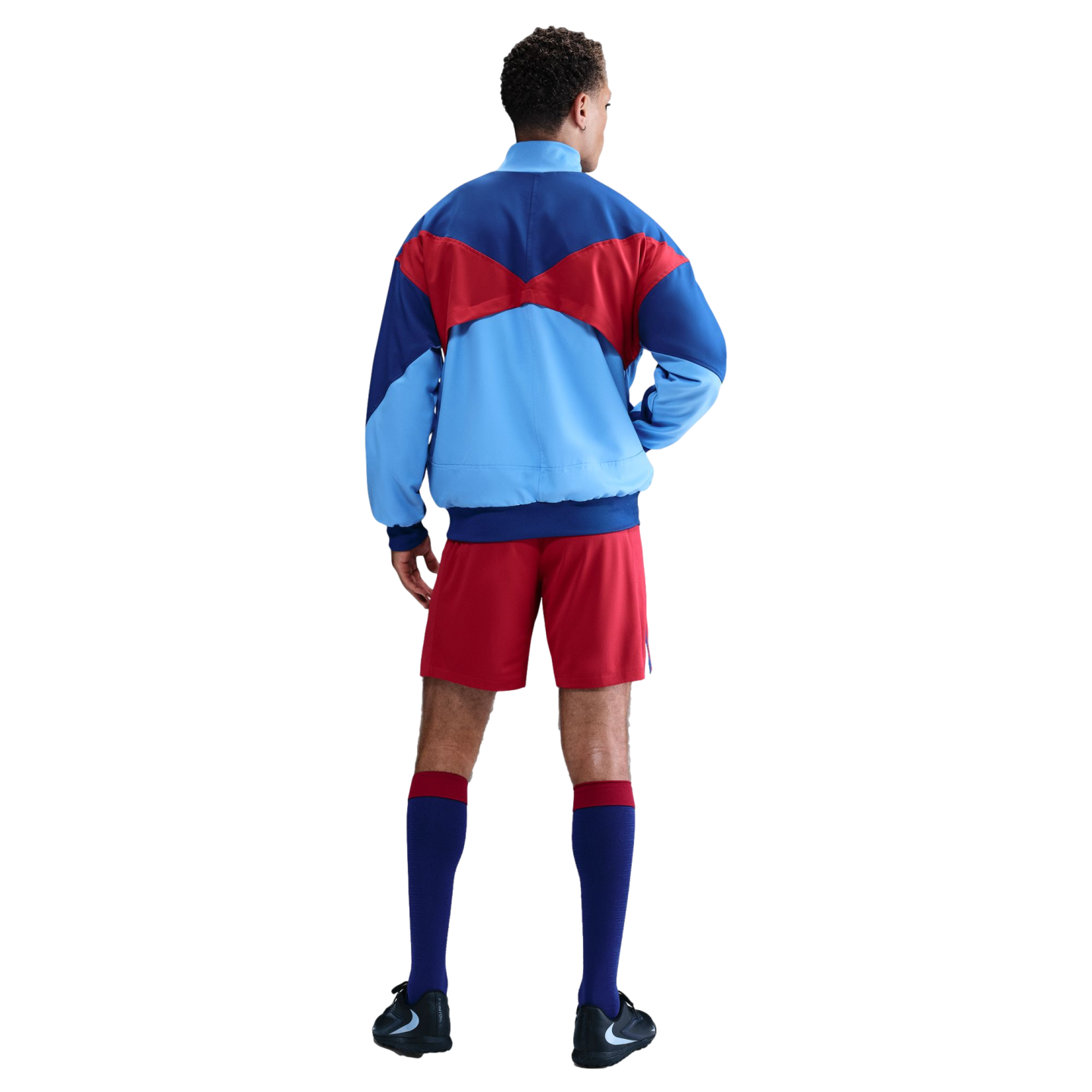 Back view of a man wearing a blue, light blue, and red color-blocked jacket, red shorts, and navy socks with red trim against a black background.