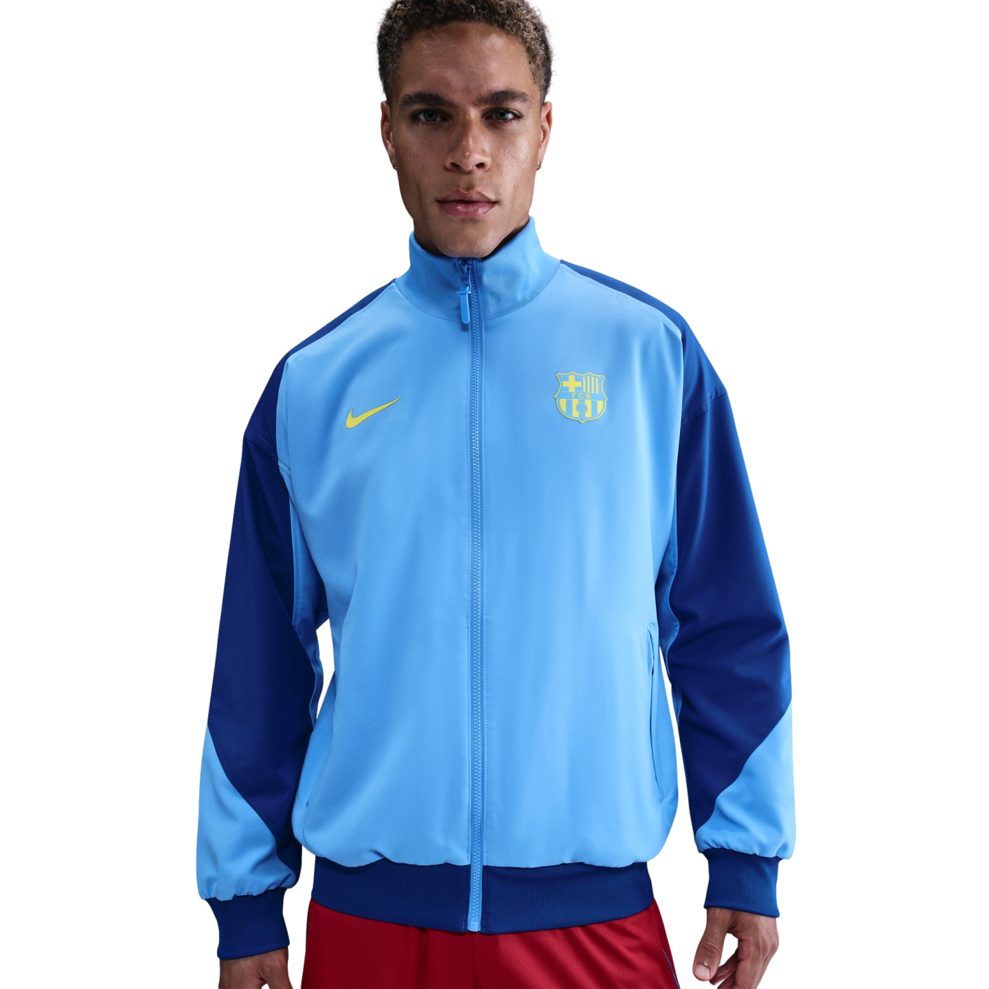 A man models a light and dark blue Nike FC Barcelona track jacket with a yellow club crest, posed against a solid black background.