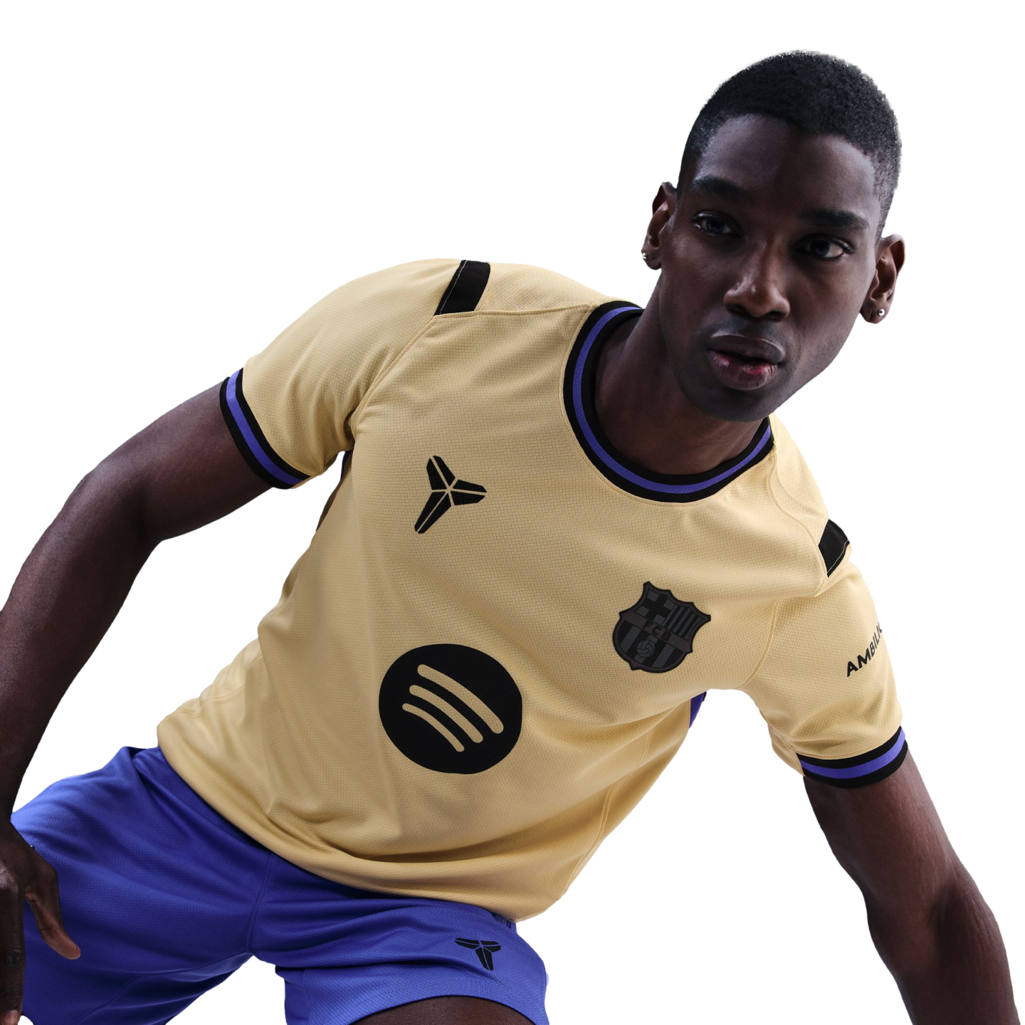 A man in a tan FC Barcelona football jersey featuring Kobe Bryant’s "Sheath" logo and a Spotify logo, wearing blue shorts against a black background.