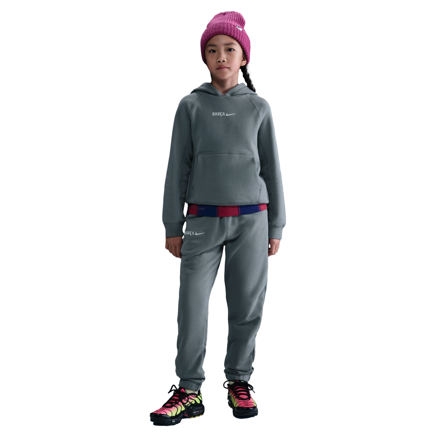 Child in a gray Nike FC Barcelona tracksuit, pink beanie, and colorful sneakers against a black background.