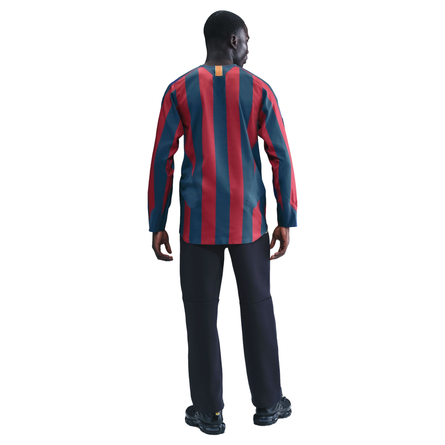 Back view of a man with dark skin wearing a long-sleeved shirt with vertical dark red and blue stripes, black trousers, and black sneakers, standing against a solid black background.