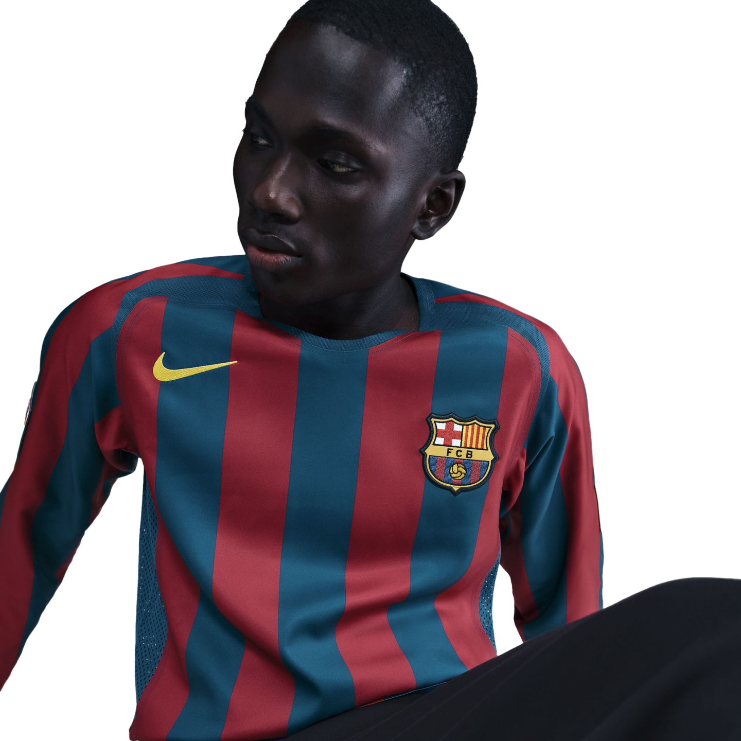 A young Black man in profile wearing a long-sleeved FC Barcelona jersey with red and blue vertical stripes, featuring a yellow Nike swoosh and the club's crest. He looks off-camera against a solid black background.