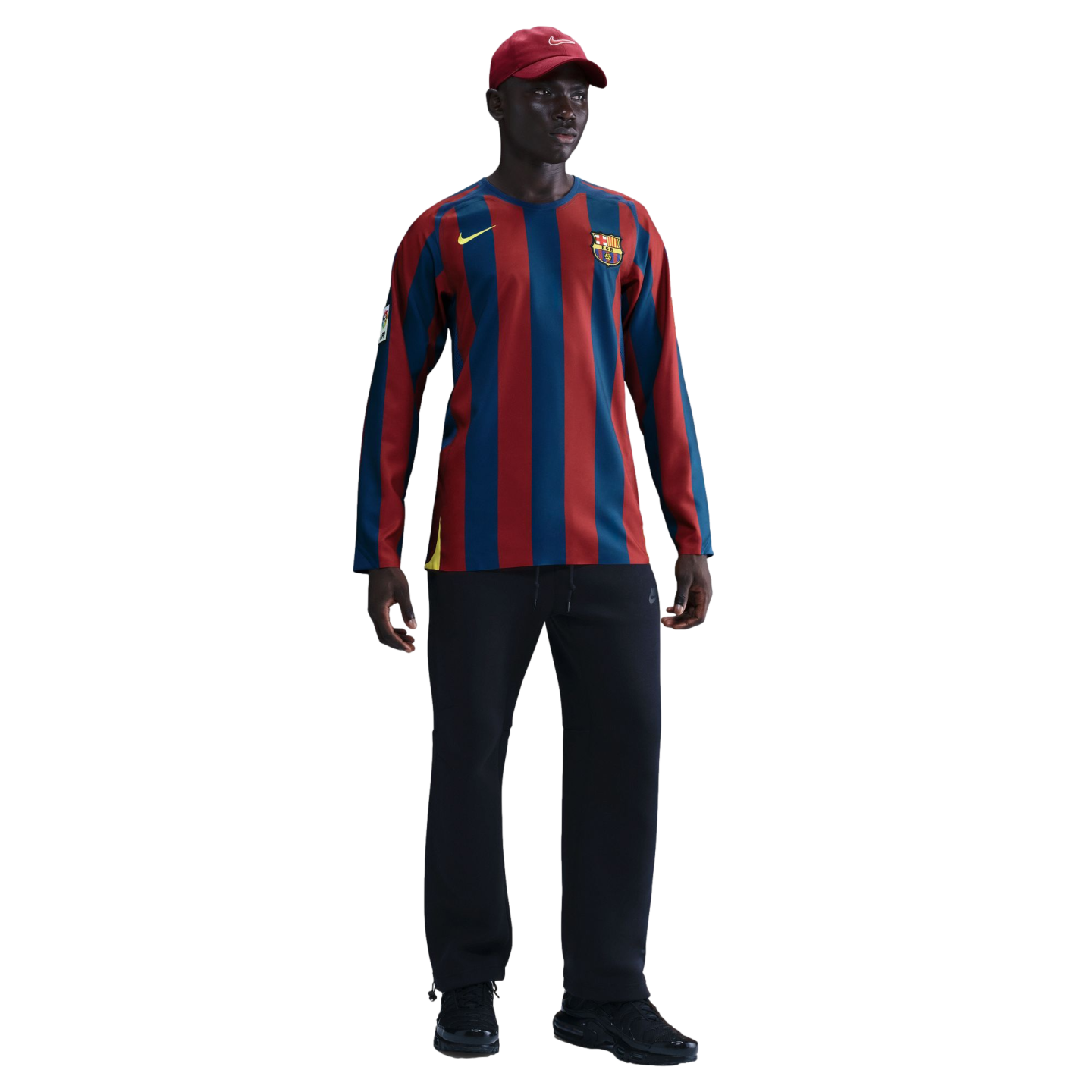 A man wearing a red cap, long-sleeved FC Barcelona striped jersey, black pants, and black sneakers, standing against a solid black background.