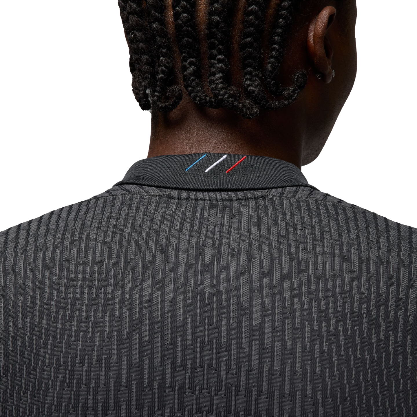 Rear view of a person with braided hair wearing a dark grey textured shirt with a small blue, white, and red tricolor detail on the back of the collar.