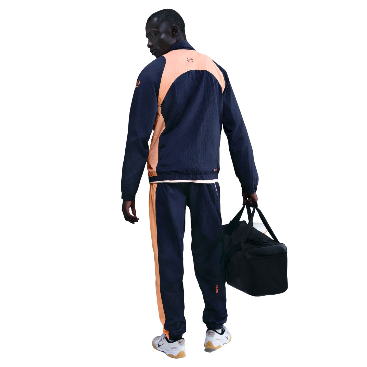 A man in a navy blue and peach tracksuit, seen from behind, holding a black gym bag against a black background.