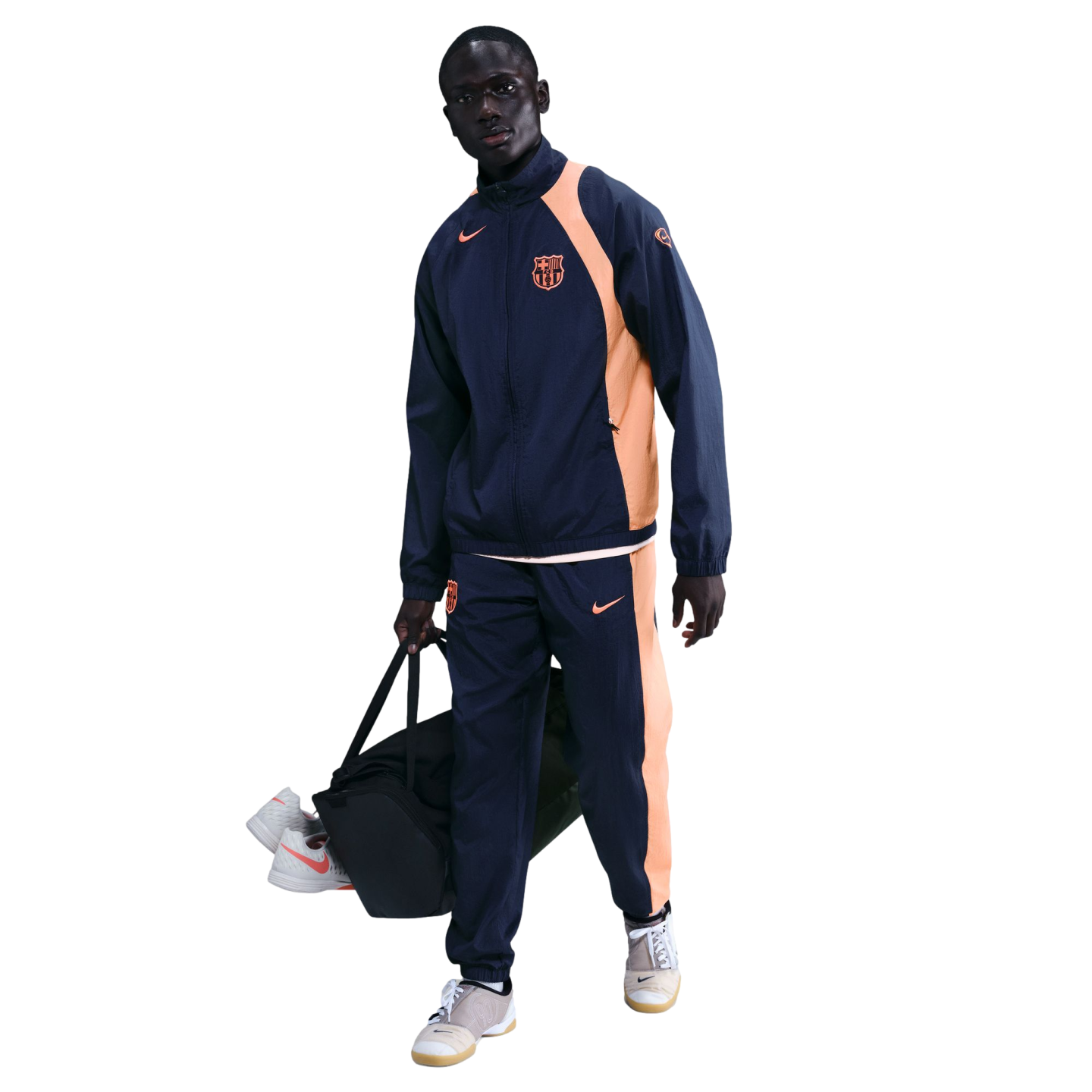 A man wearing a navy and orange Nike FC Barcelona tracksuit carries a black duffel bag against a solid black background.