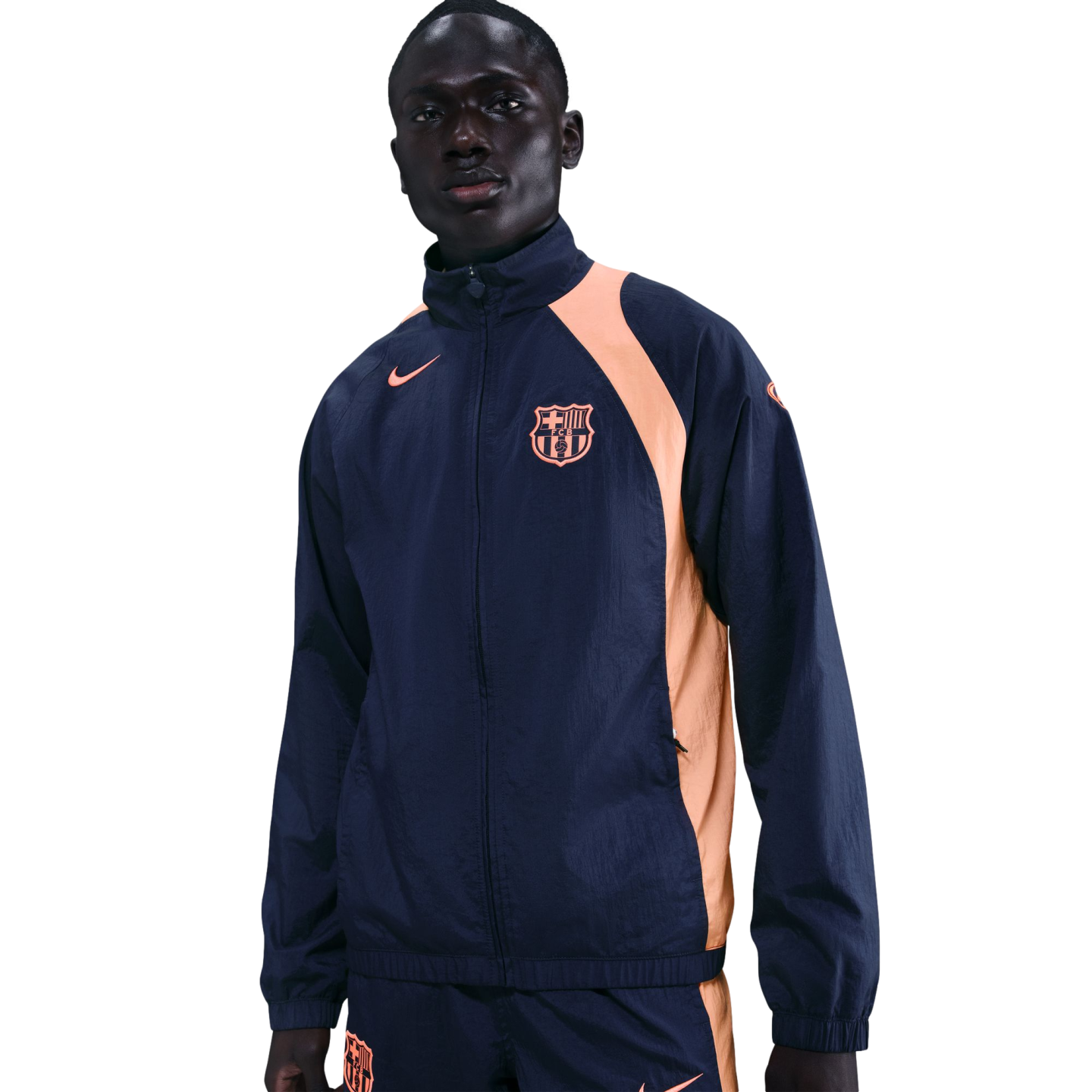 A man wearing a navy blue and peach Nike FC Barcelona windbreaker tracksuit against a black background.