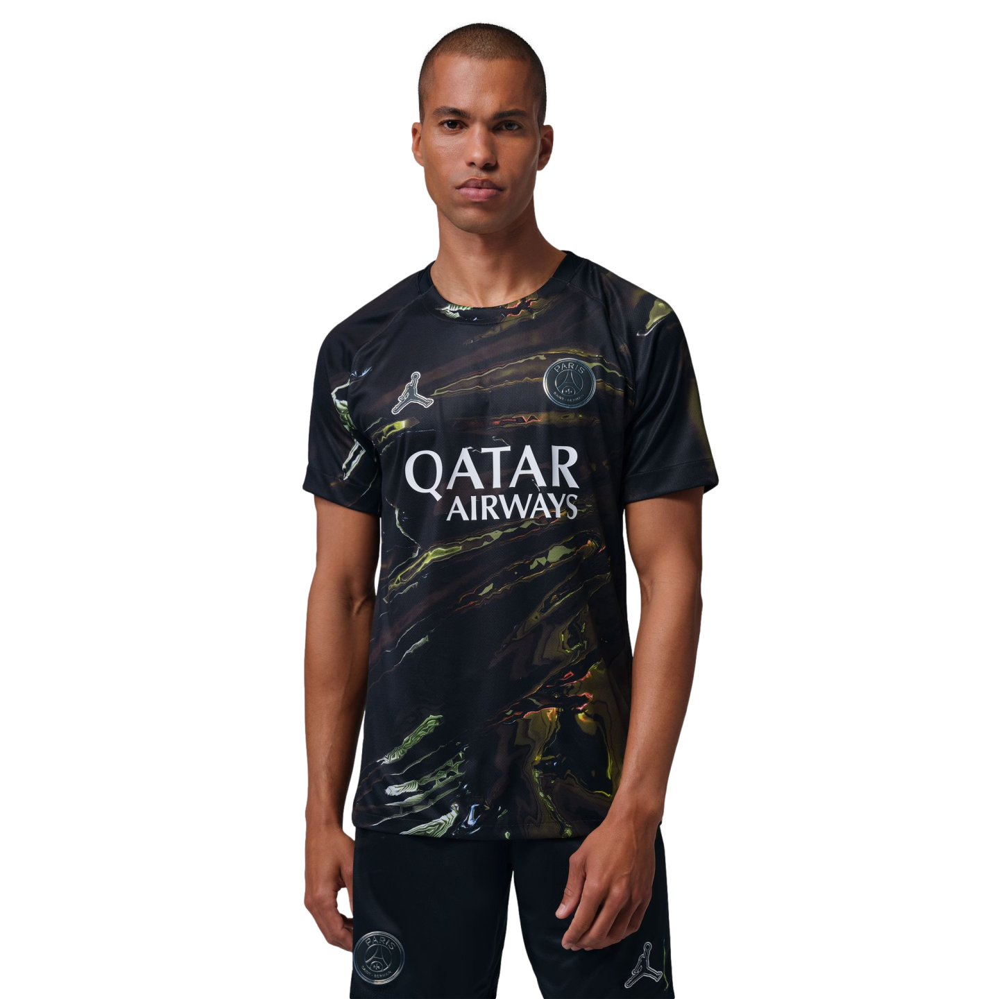 A man with a buzz cut models a black Paris Saint-Germain jersey and shorts featuring an iridescent marbled pattern, the Qatar Airways sponsor logo, and Jordan Brand's Jumpman logo, set against a solid black background.