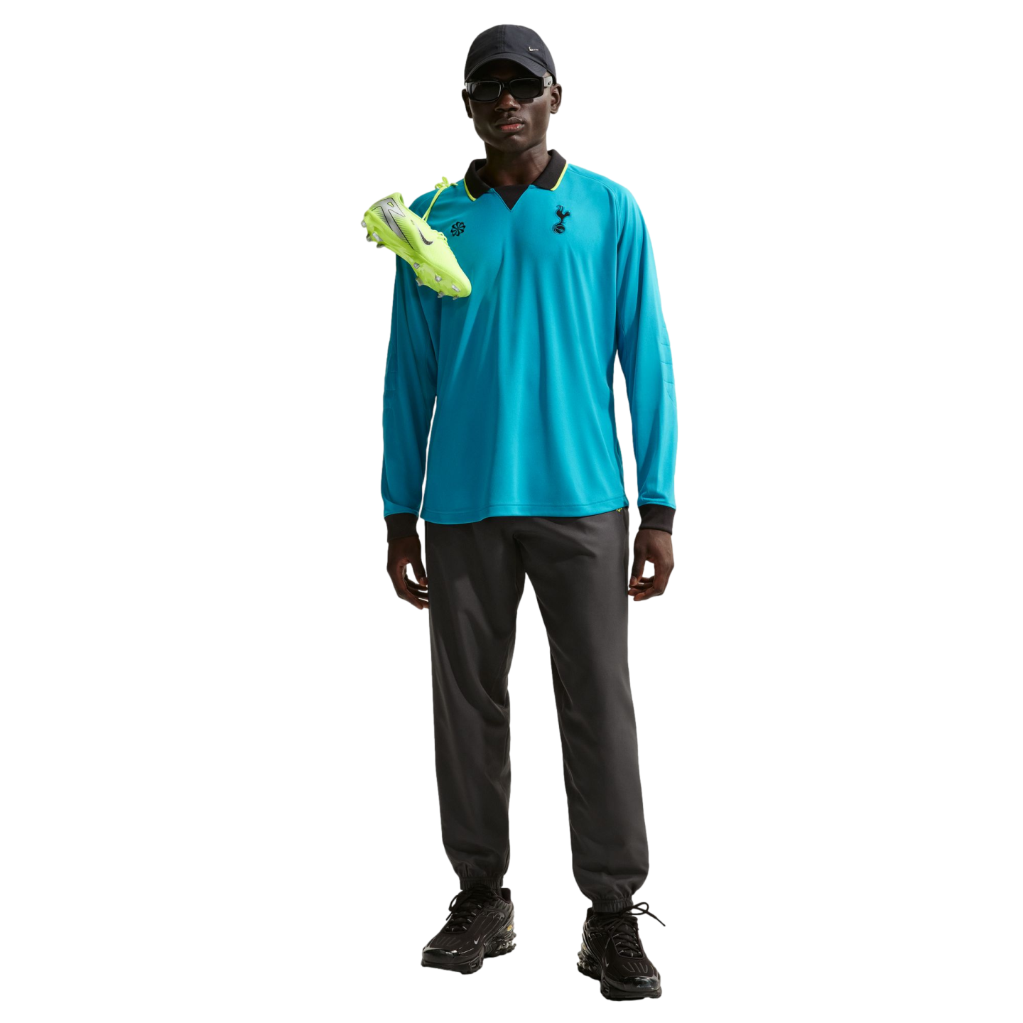 A person in a blue Tottenham long-sleeve polo, grey joggers, black cap, and sunglasses with neon green soccer cleats draped over their shoulder.