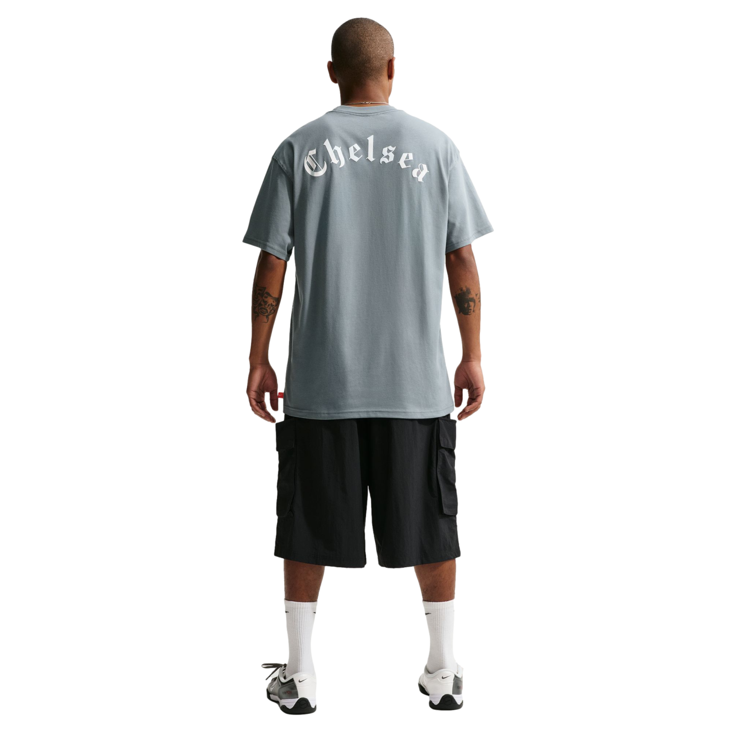 Back view of a man wearing a grey t-shirt with "Chelsea" in white gothic font, black cargo shorts, white socks, and sneakers against a black background.