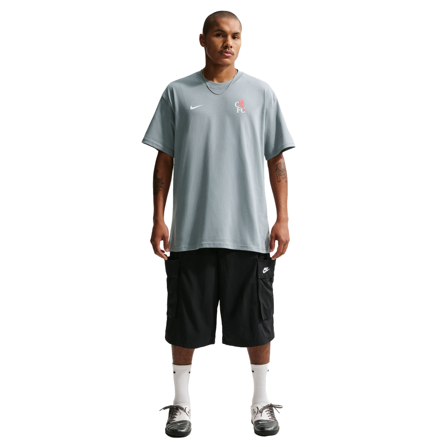 Full-length shot of a man in a grey Nike t-shirt, black cargo shorts, white socks, and grey shoes against a black background. He has a necklace and arm tattoos.