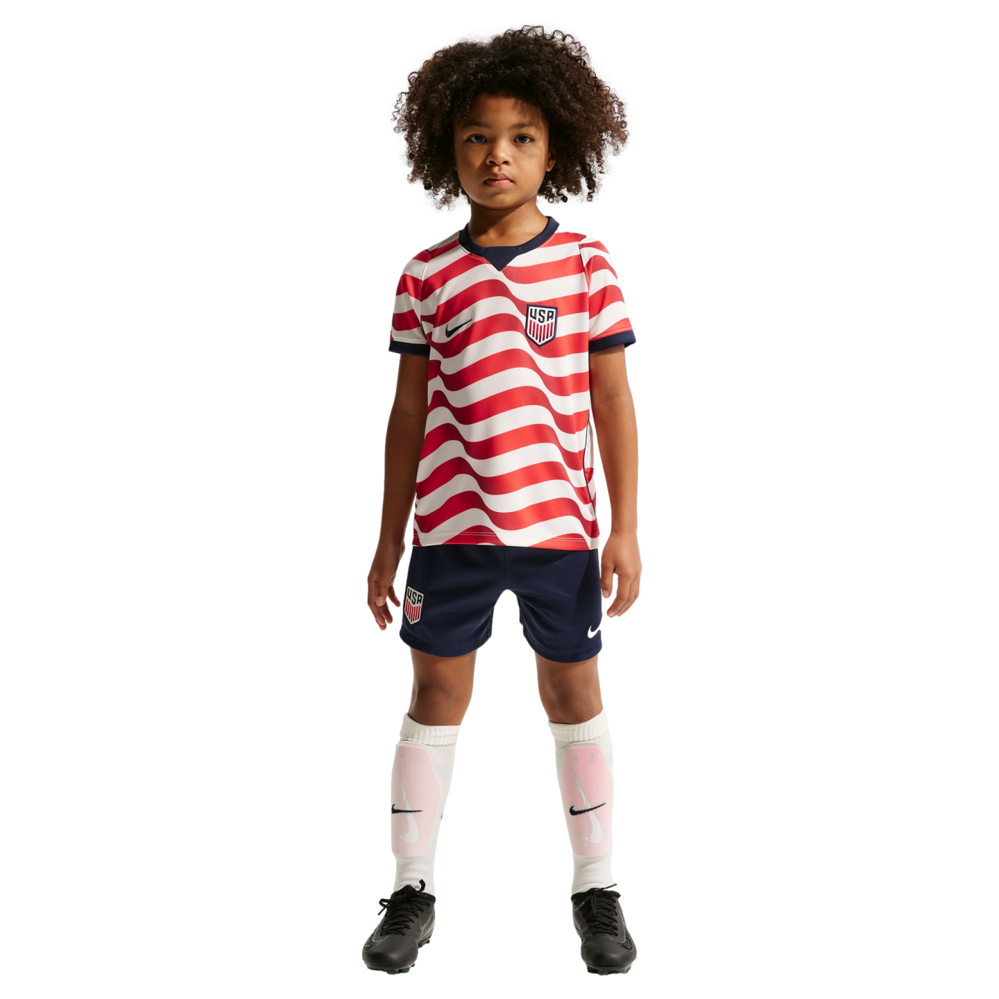 Young child with curly hair wearing a red and white wavy-striped US soccer jersey, navy shorts, and white socks against a solid black background.