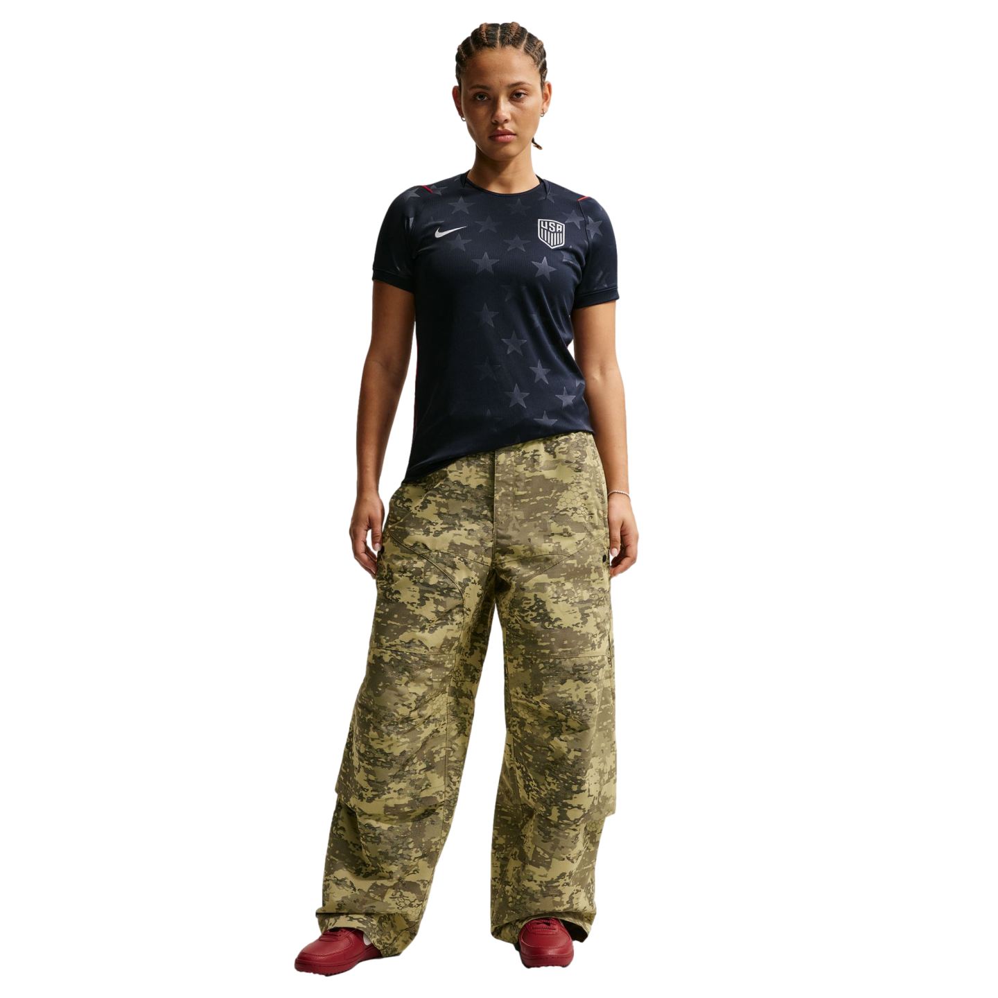 A woman stands against a black background wearing a navy USA soccer jersey, oversized camouflage cargo pants, and red sneakers.