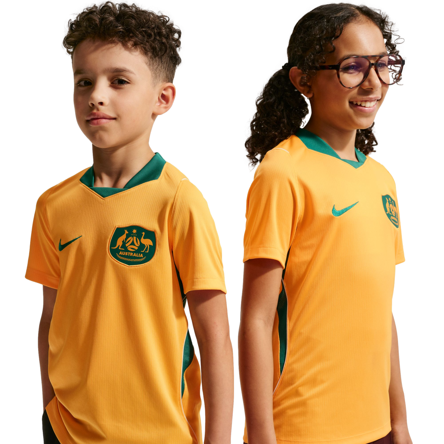 A boy and a girl pose side-by-side against a black background wearing gold Nike Australia national soccer team jerseys with green trim. The boy looks towards the camera while the girl, wearing glasses, smiles and looks off to the right.