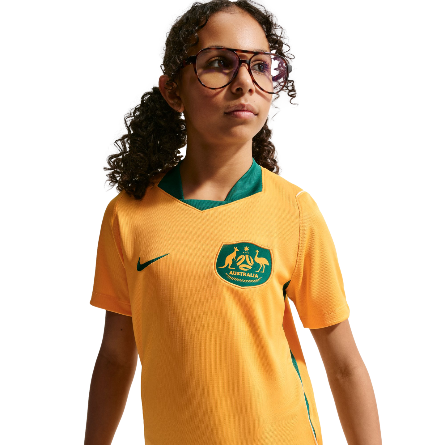 A waist-up portrait of a young girl with curly dark hair and glasses, wearing a yellow and green Australian national soccer team jersey against a black background.