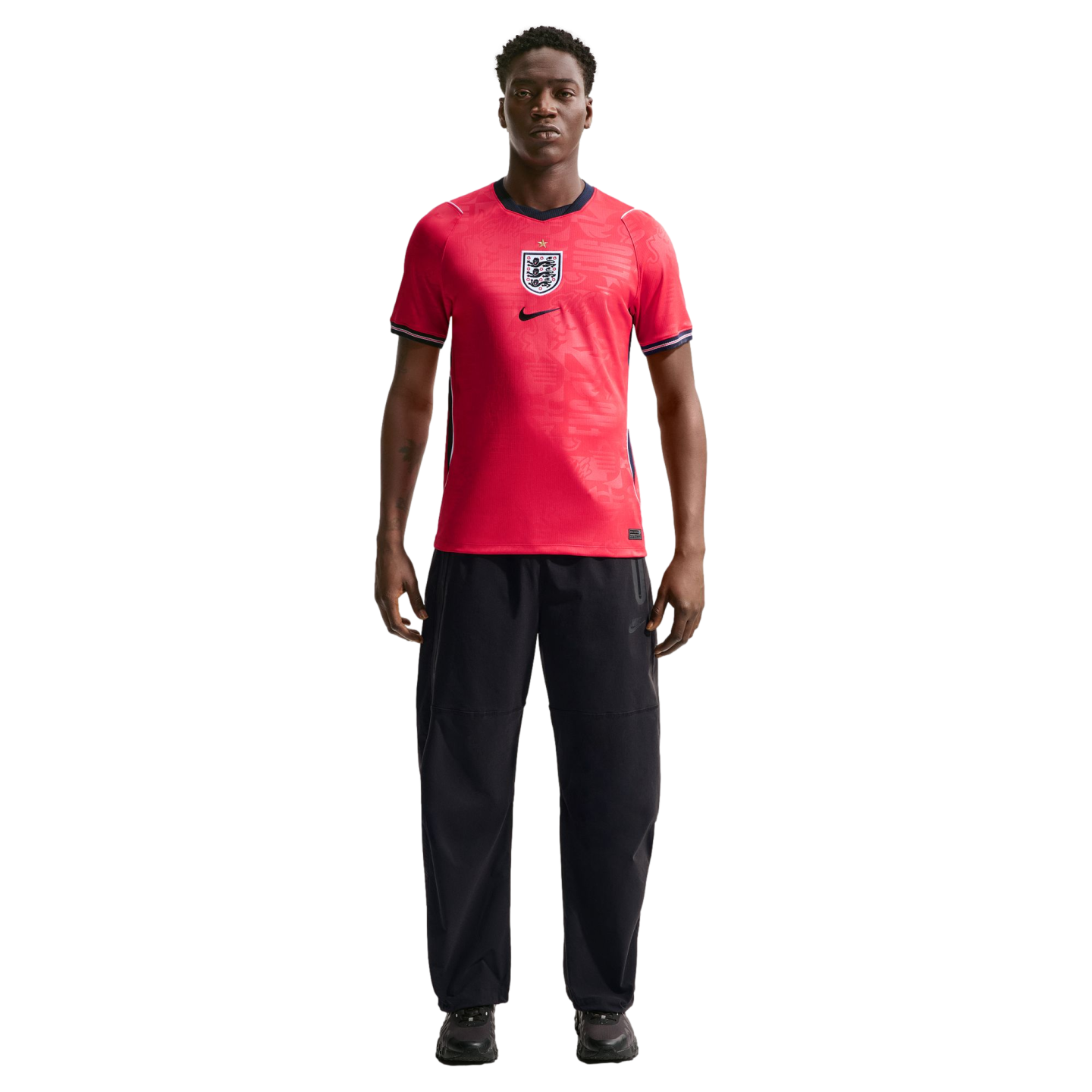 A man stands in a red England national football team jersey and black trousers against a plain black background.