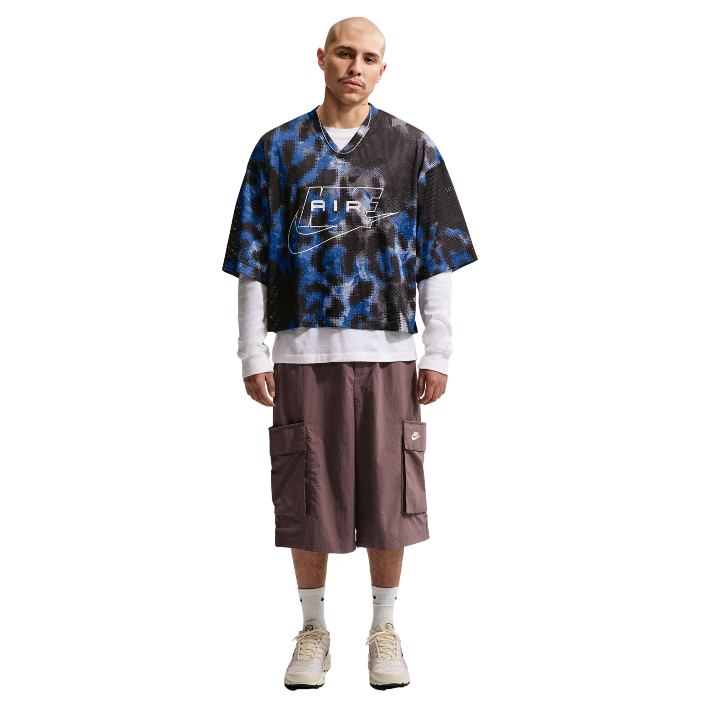 A man wears an oversized blue and black patterned Nike jersey, white long-sleeve shirt, baggy brown cargo shorts, and cream sneakers against a white background.