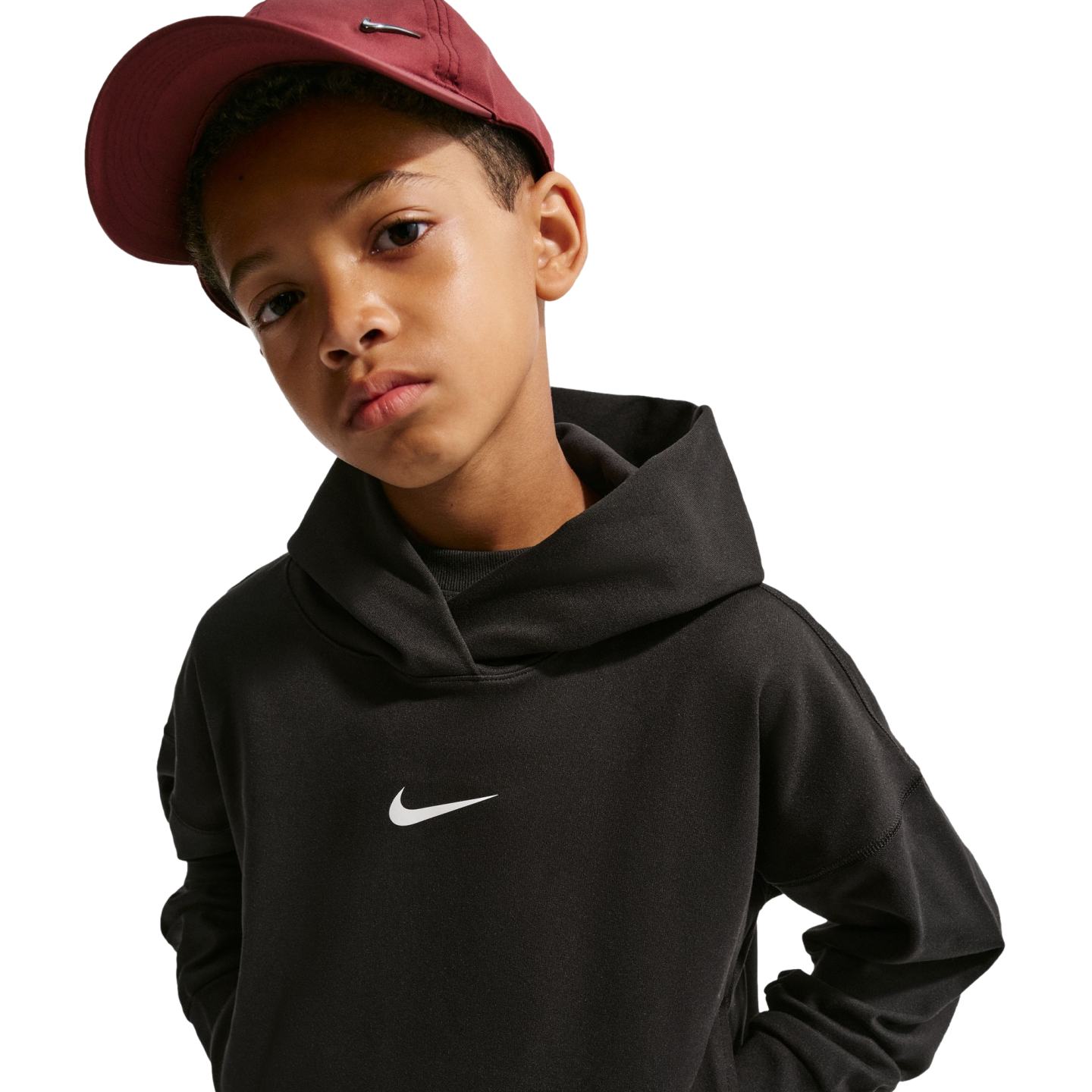 A boy wearing a black Nike hoodie and a maroon Nike baseball cap looks at the camera against a black background.