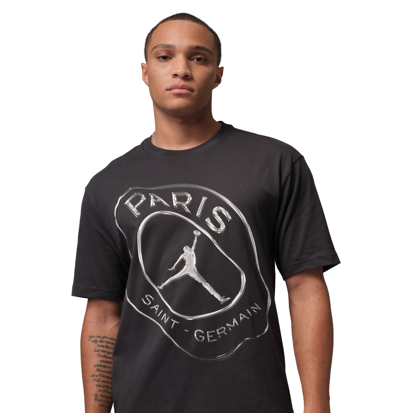 A man wearing a black t-shirt with a metallic silver graphic of the Paris Saint-Germain logo and Jordan Brand Jumpman on a black background.