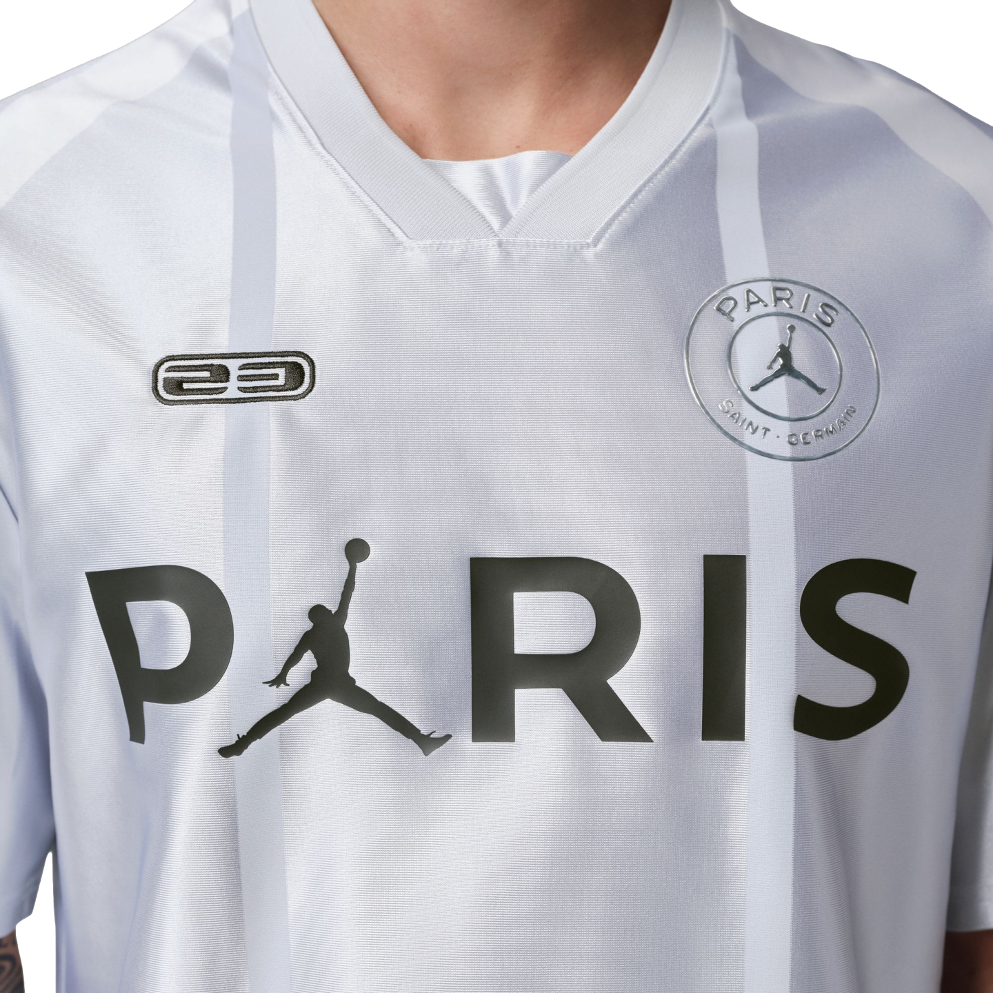 Close-up of a white PSG jersey featuring a large black 'PARIS' graphic with a Jumpman logo replacing the letter 'A', faint grey stripes, and team crest.