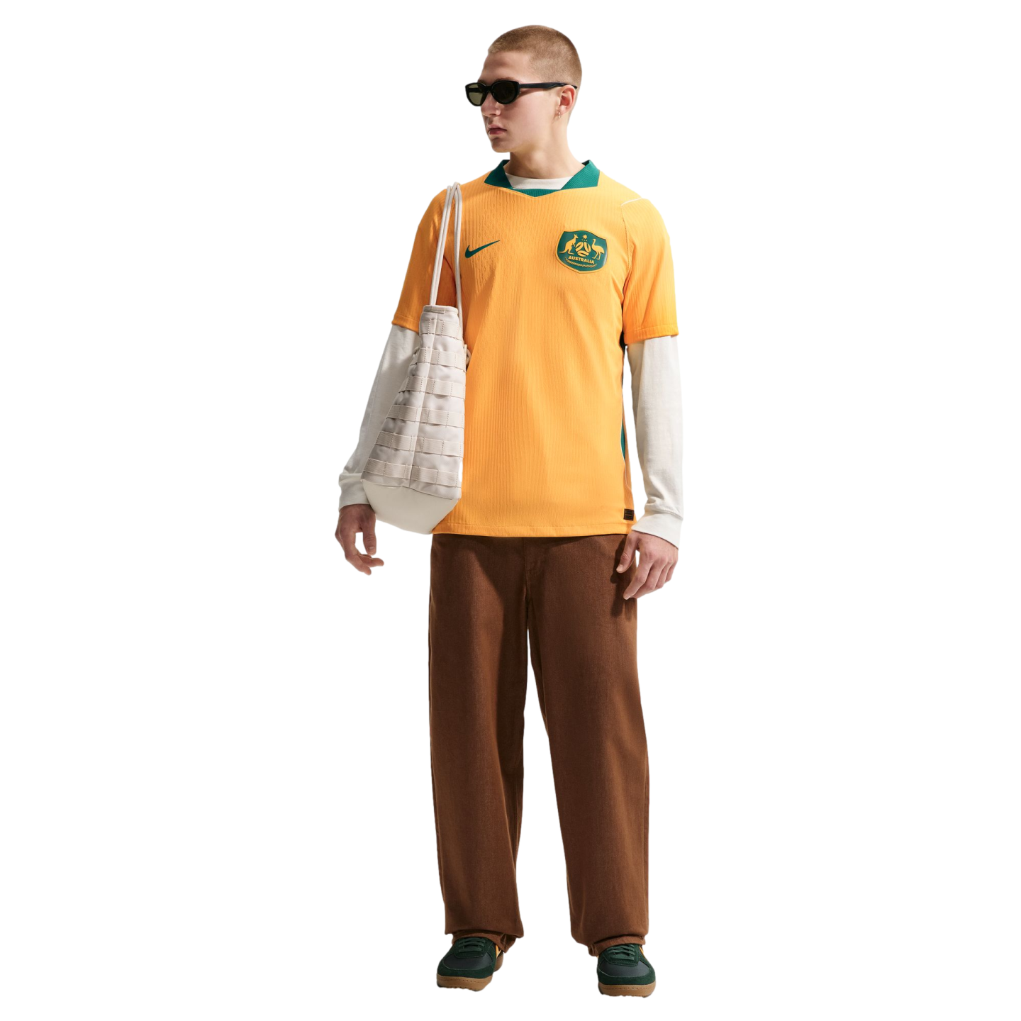 A man in a yellow Australia soccer jersey, white long-sleeved shirt, brown pants, and green sneakers carries a white tote bag against a black background.