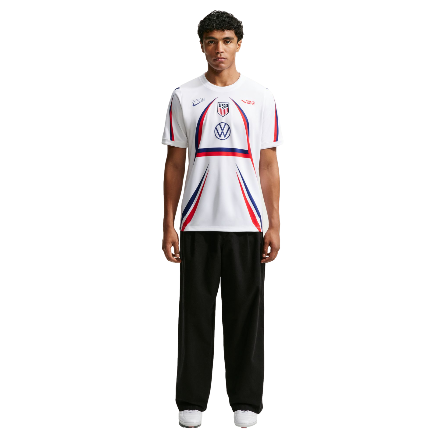 A man stands wearing a white USA soccer jersey with red and blue accents, black wide-leg trousers, and white sneakers against a plain white background.