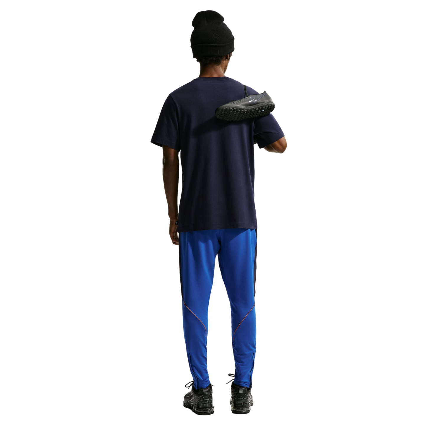 Rear view of a person wearing a navy t-shirt, bright blue track pants with orange trim, and a black beanie. They have a black Nike soccer cleat slung over their shoulder against a solid black background.
