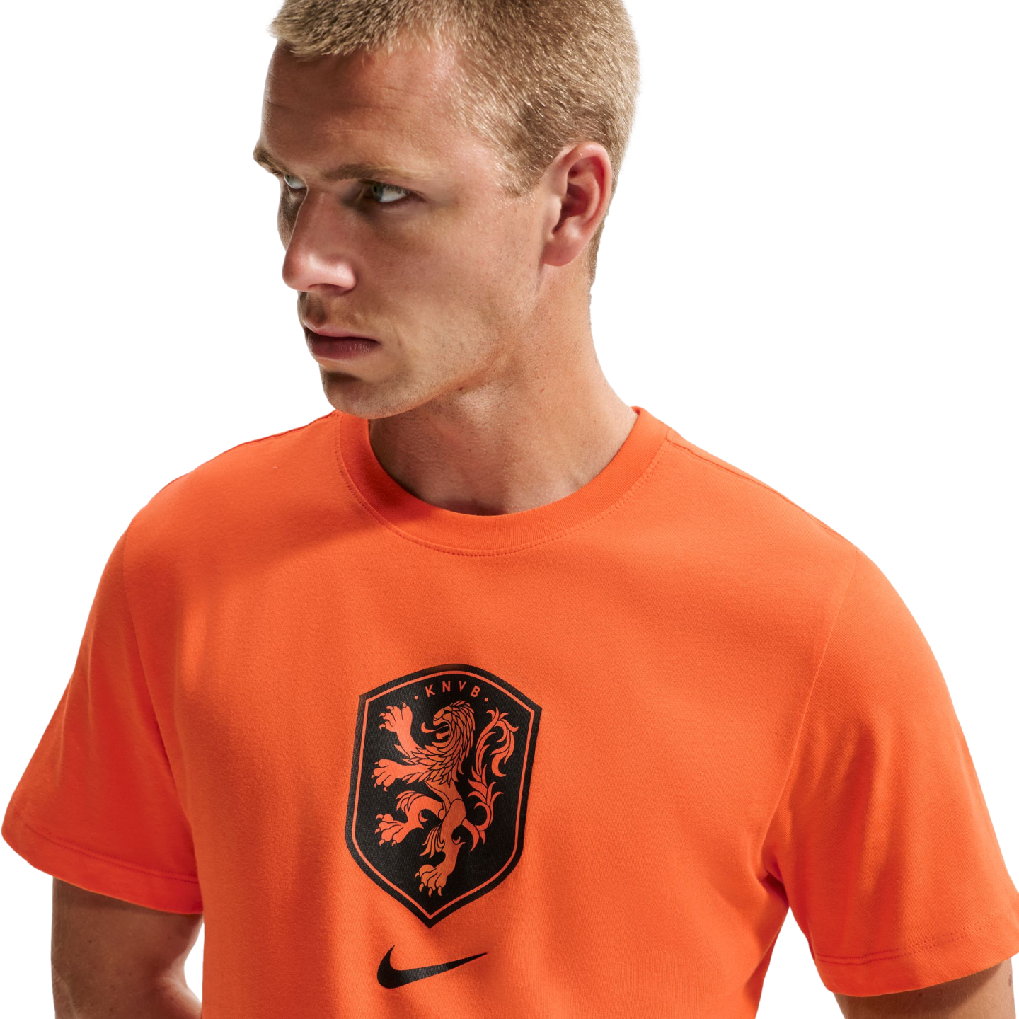 A man with short blond hair wearing a bright orange Nike t-shirt featuring a black KNVB lion crest. He is looking off to the side against a solid black background.