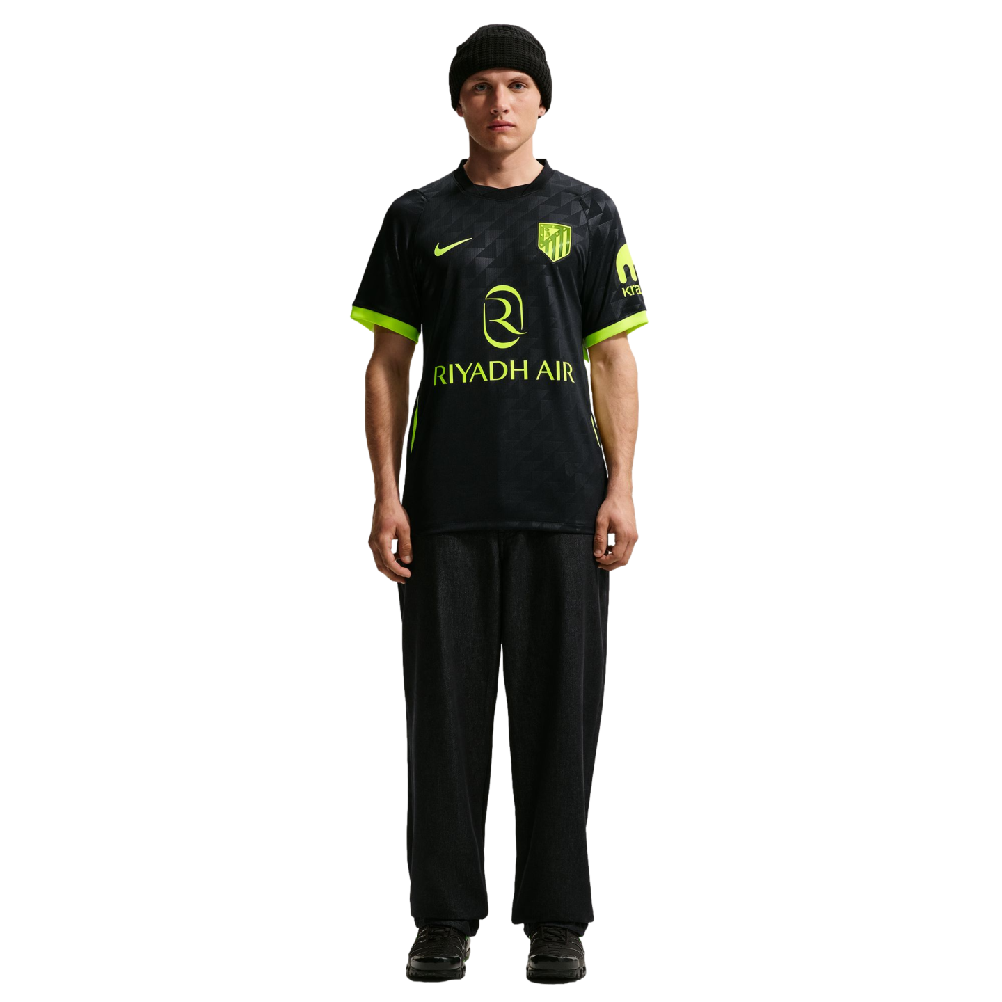 Man in a black Nike Atletico Madrid jersey with neon accents and "RIYADH AIR" sponsor, paired with black pants and a beanie, standing against a white background.