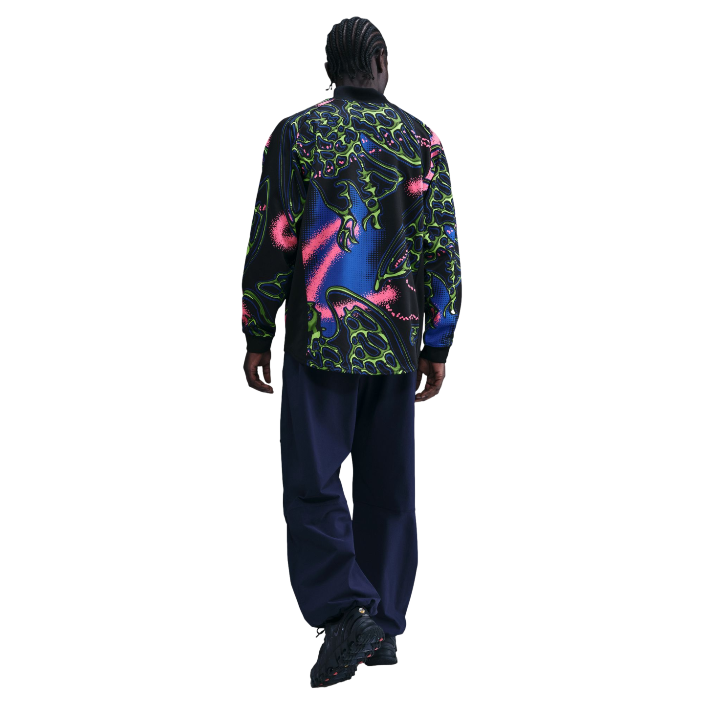 Rear view of a person with cornrows walking away, wearing a neon-patterned black shirt, navy trousers, and chunky black sneakers against a black background.