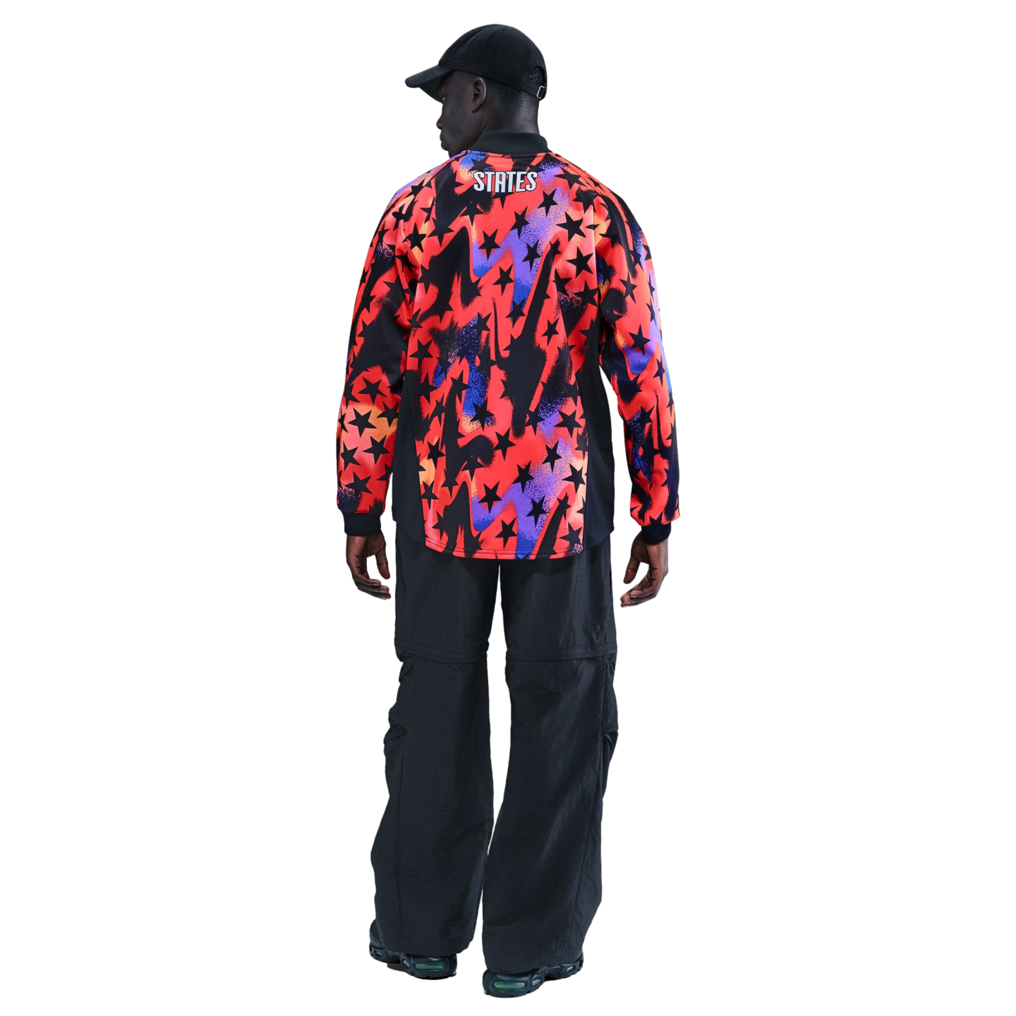 Rear view of a man wearing a black cap, a multi-colored star-patterned shirt with "STATES" on the back, and baggy black pants against a black background.