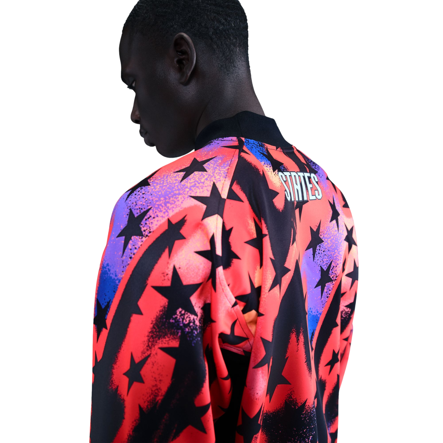 A man from the back wearing a vibrant jacket with black stars over red, orange, and purple gradients. The word "STATES" is printed on the upper back. Black background.