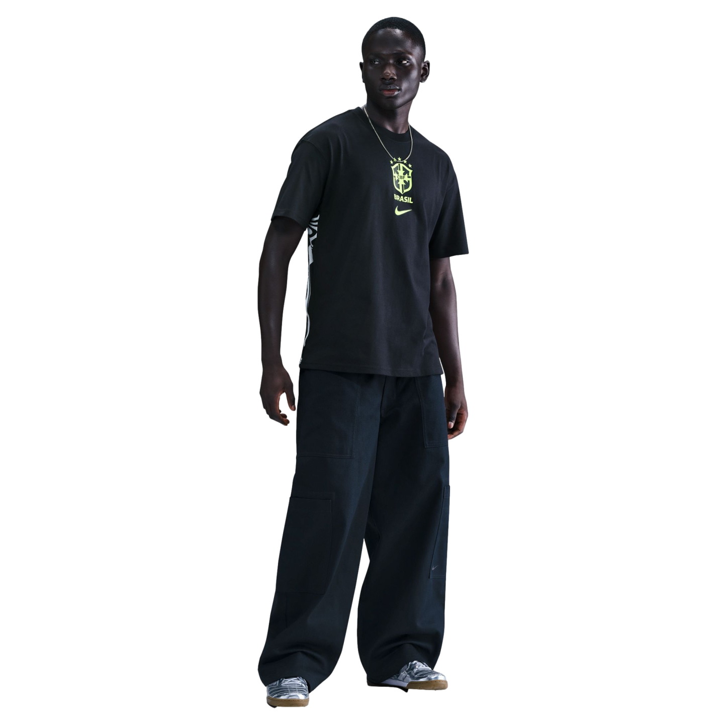A man models a black Nike t-shirt with a neon yellow Brazil crest, wide-leg black cargo pants, and silver sneakers against a black background.