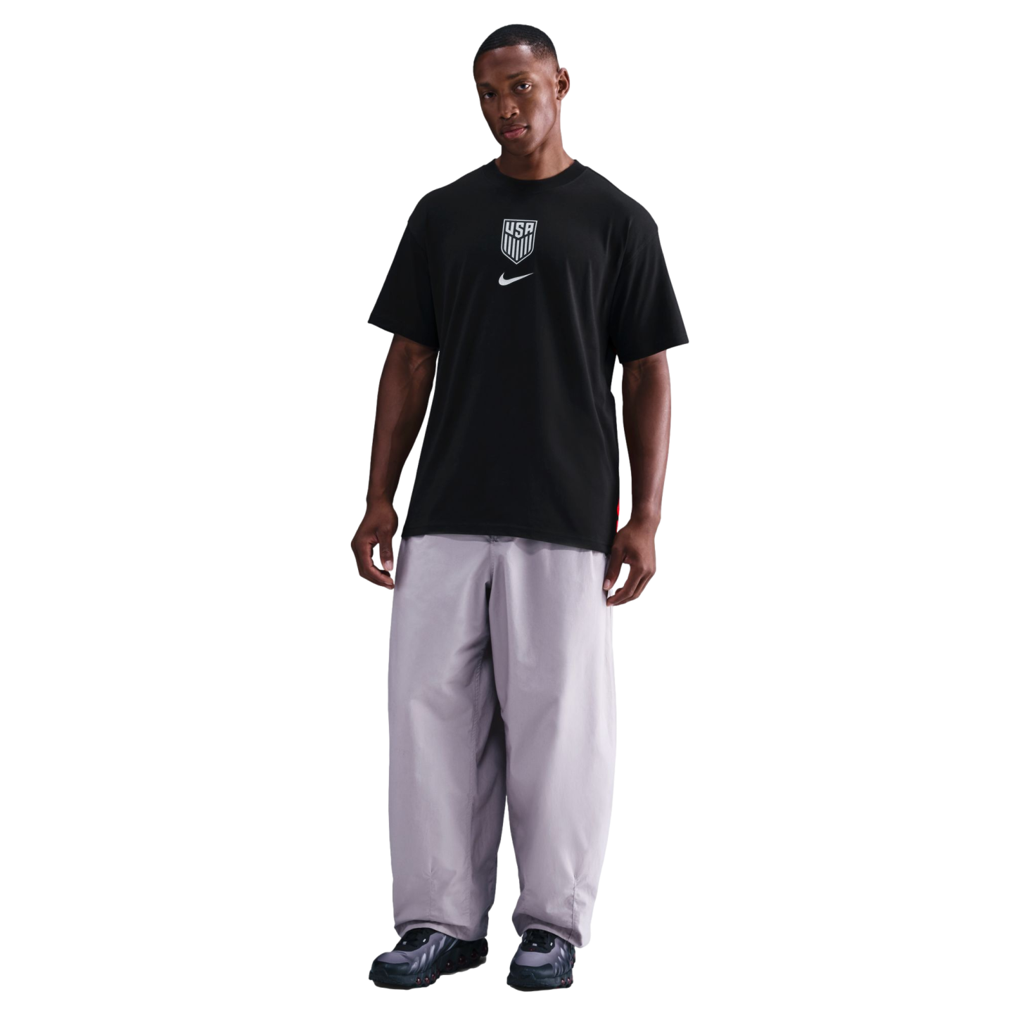 A man stands against a black background wearing a black Nike T-shirt with a USA crest and loose grey pants.