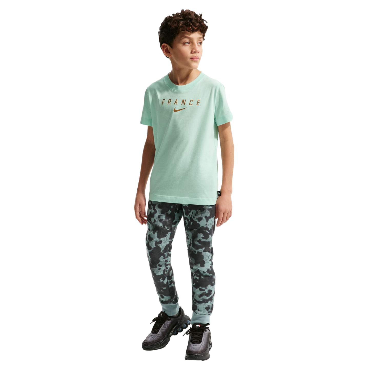 A young boy stands against a solid black background. He is wearing a light mint green short-sleeved T-shirt with the word "FRANCE" and a Nike swoosh in gold, patterned camouflage joggers, and black sneakers.