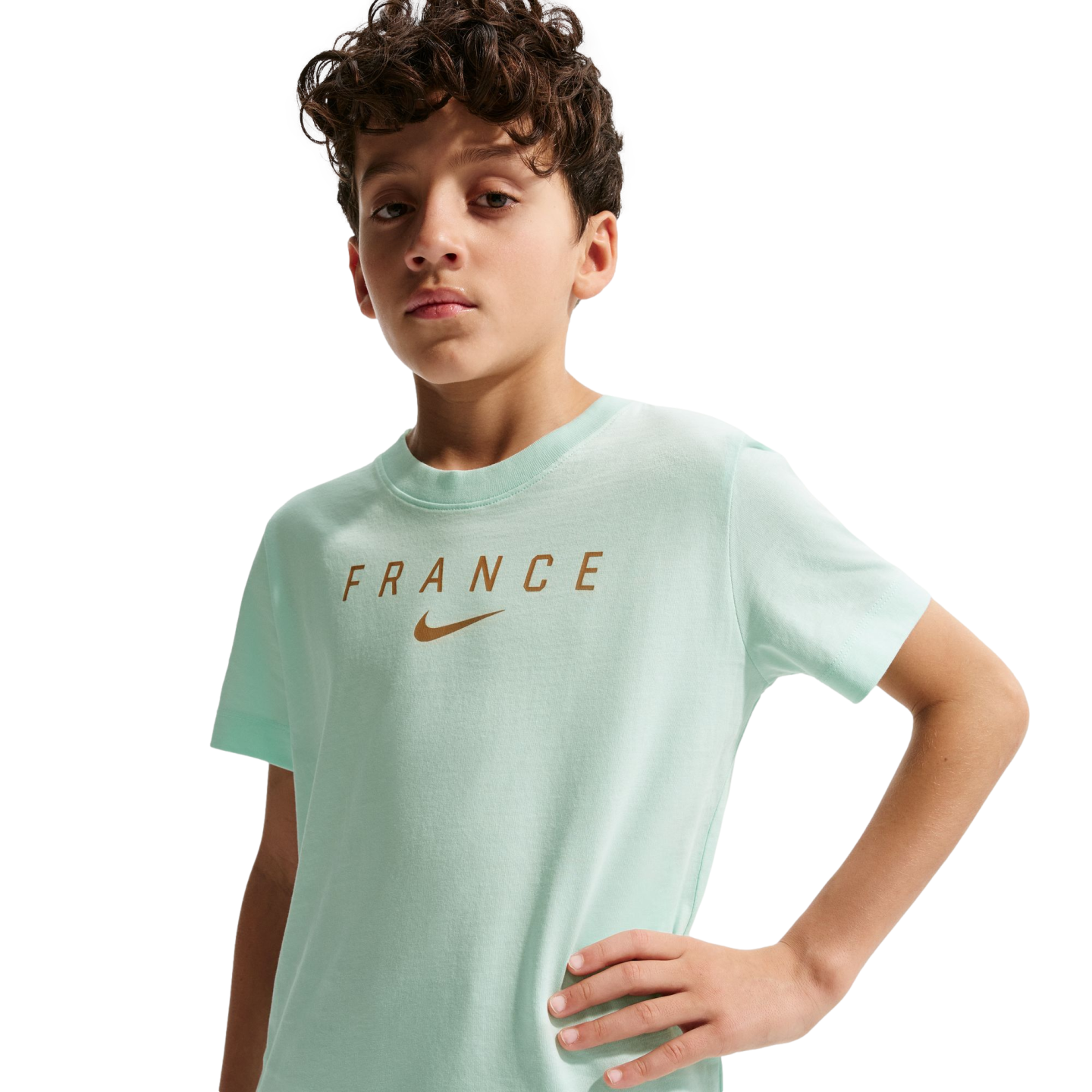 A young boy with curly brown hair wearing a light mint green t-shirt with "FRANCE" and a Nike swoosh printed in gold across the chest. He stands with one hand on his hip against a solid black background.