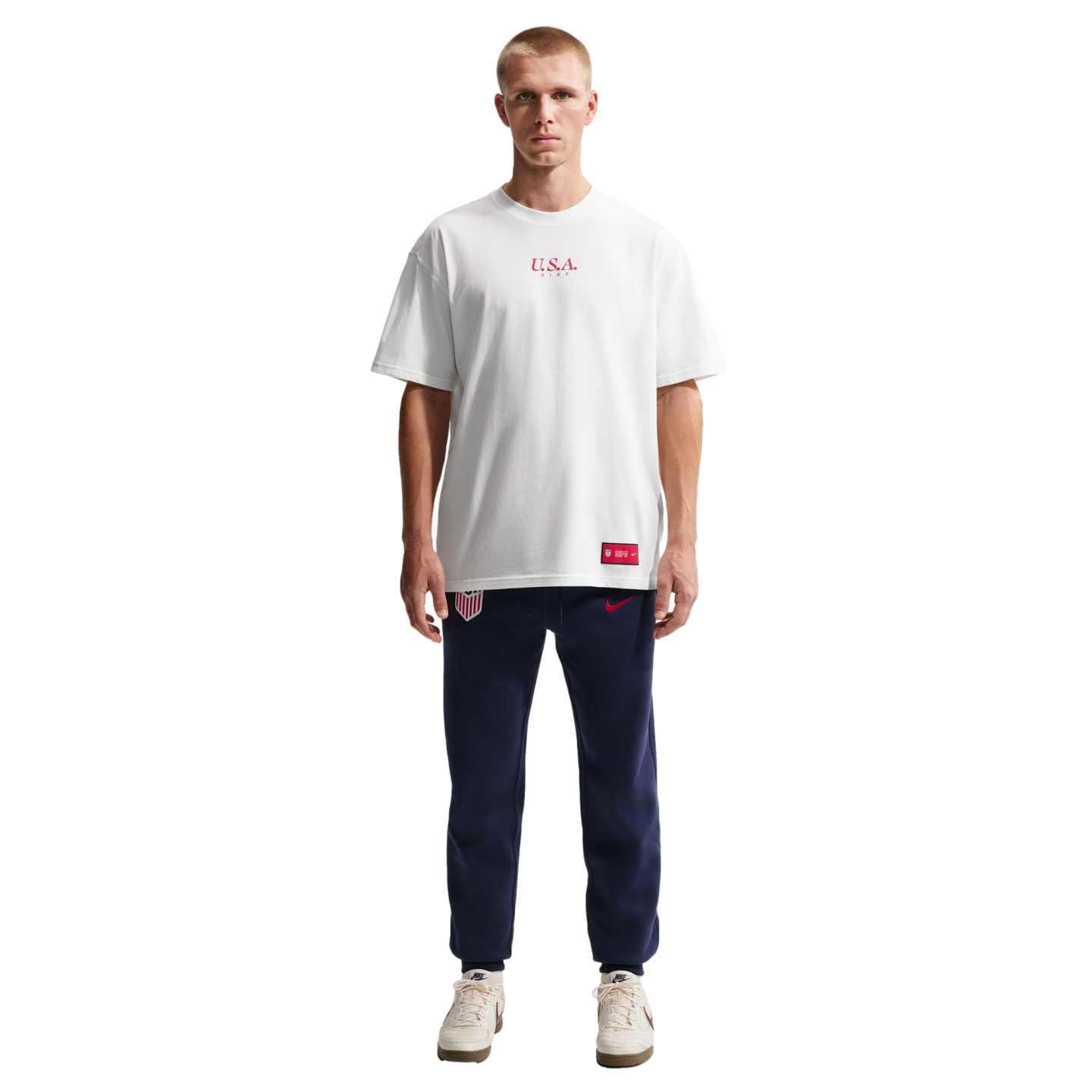A man stands against a black background wearing a white Nike U.S.A. t-shirt, navy blue USA sweatpants, and white sneakers.