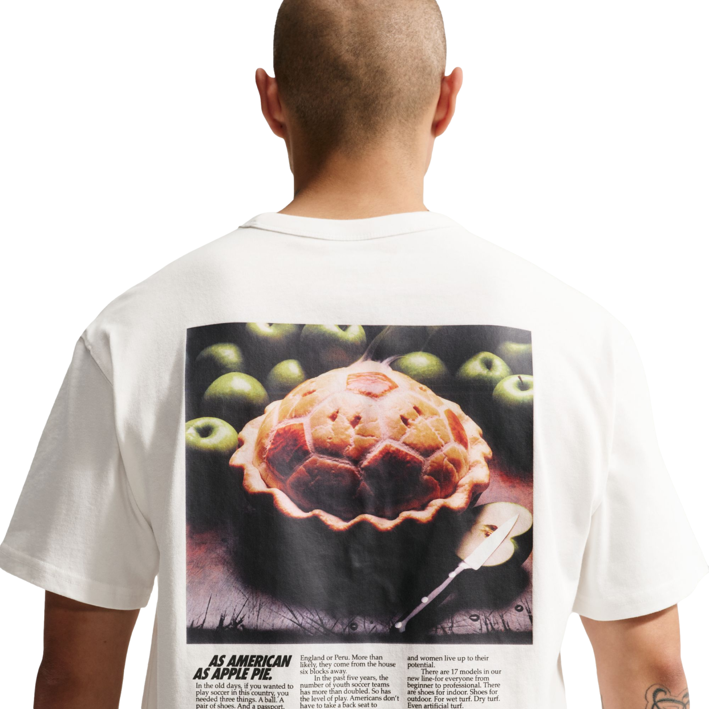 A back view of a person with a shaved head wearing a white t-shirt against a black background. The shirt features a large graphic on the back resembling a vintage magazine advertisement. The main image shows a baked pie with a soccer-ball-patterned crust 