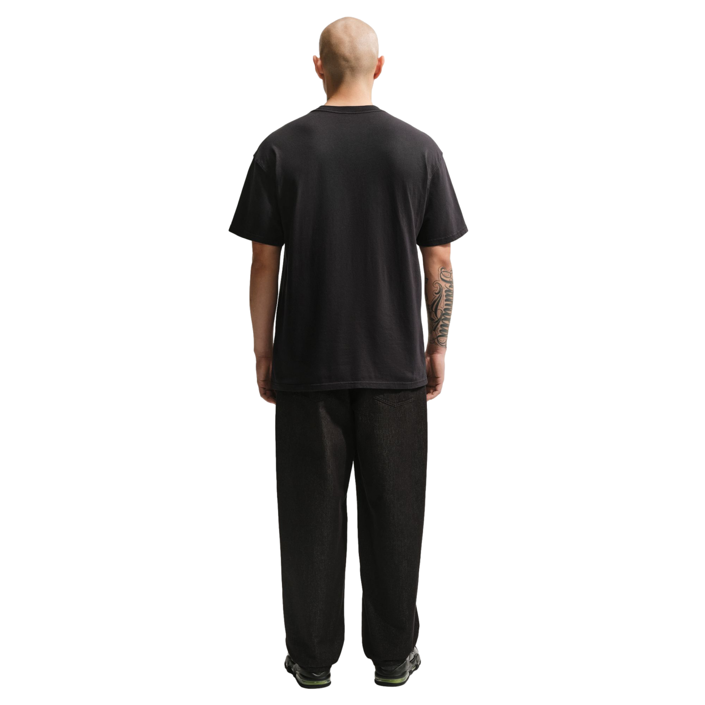 A full-length rear view of a bald man wearing a plain black t-shirt and loose-fitting black pants against a solid black background. He has a large tattoo on his right forearm.