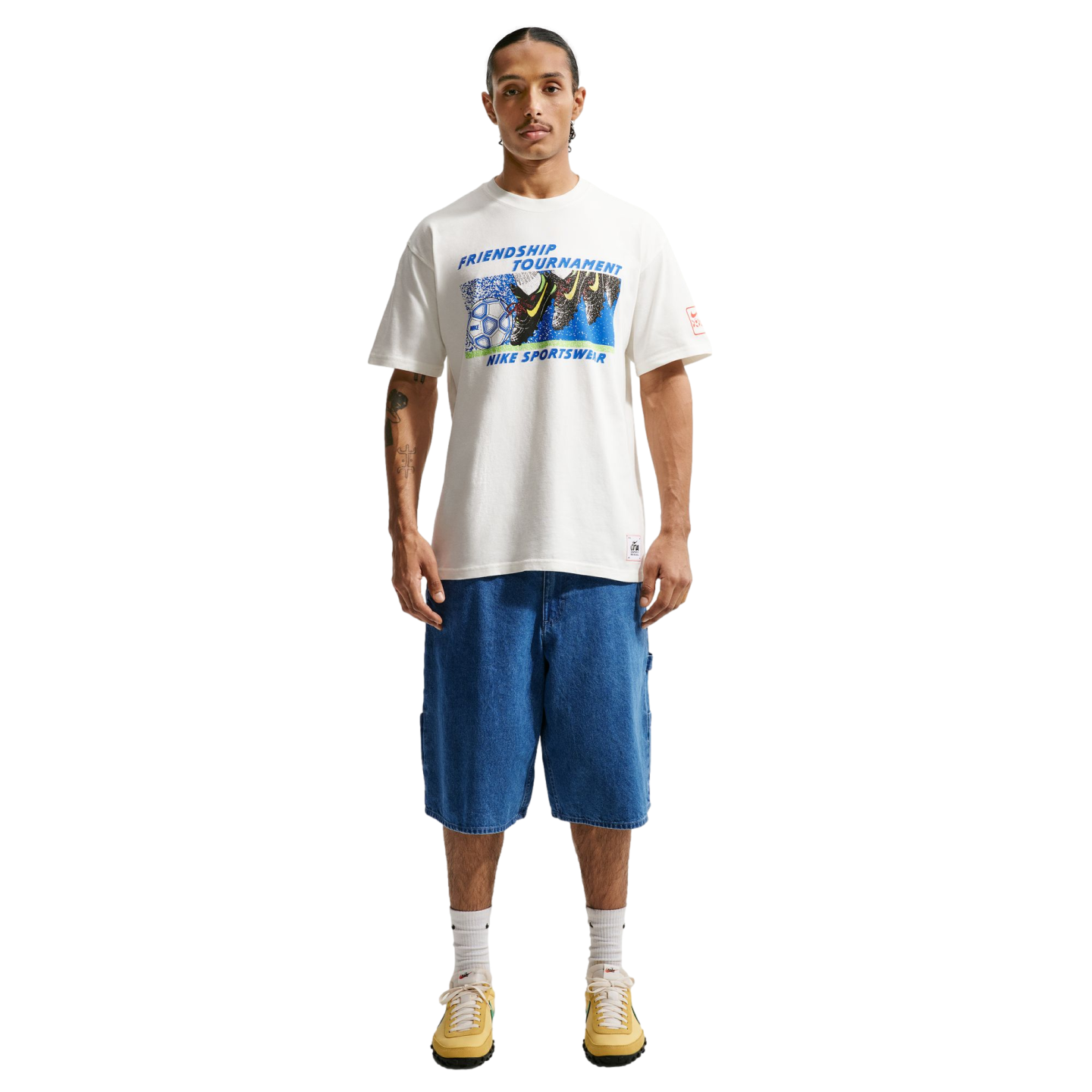 A person stands against a black background wearing a white "Friendship Tournament" Nike graphic t-shirt, knee-length blue denim shorts, white socks, and yellow sneakers.