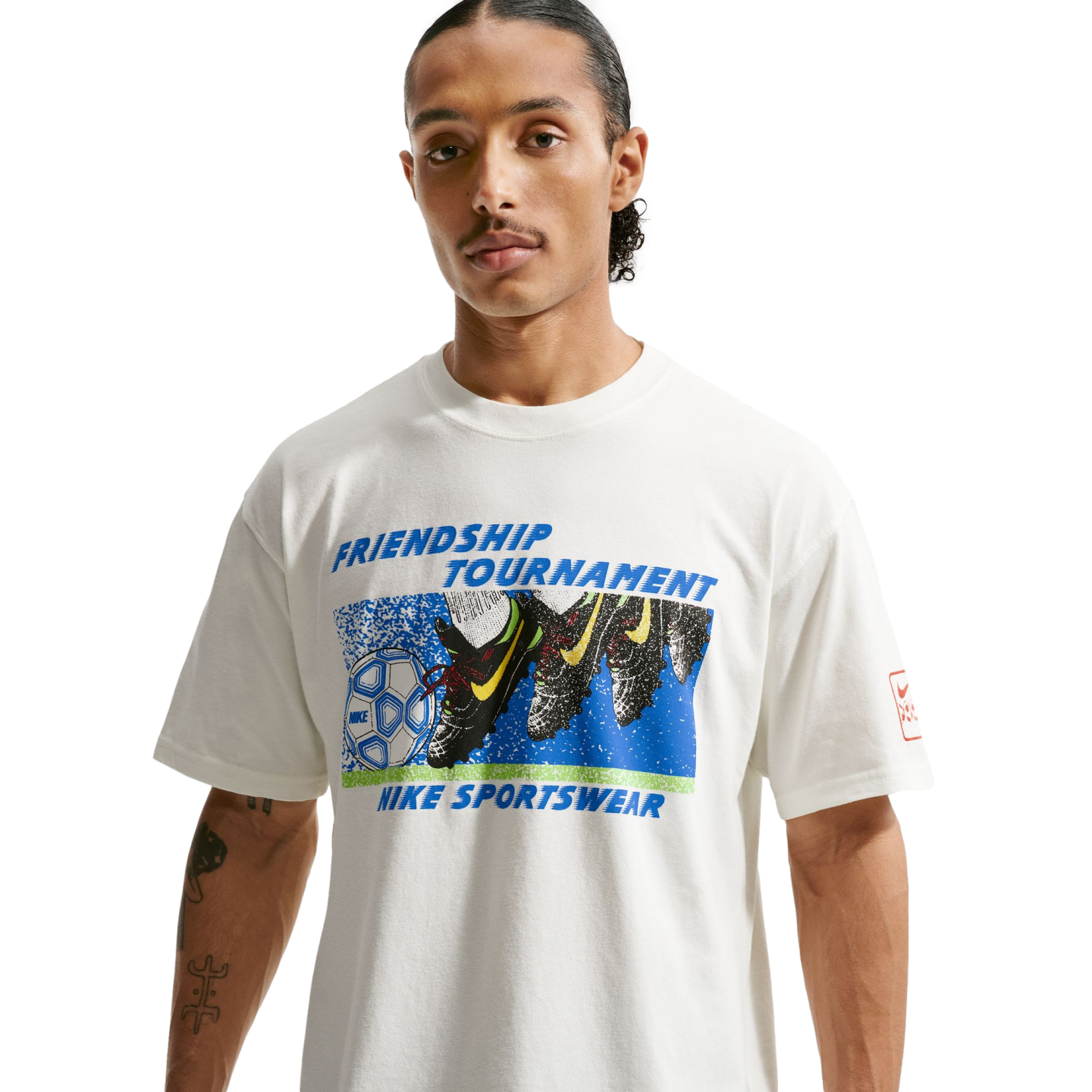 A man with a thin mustache and arm tattoos wears a white t-shirt featuring a colorful graphic of soccer cleats and a ball with the text "Friendship Tournament" and "Nike Sportswear" in blue. He is posed against a solid black background.