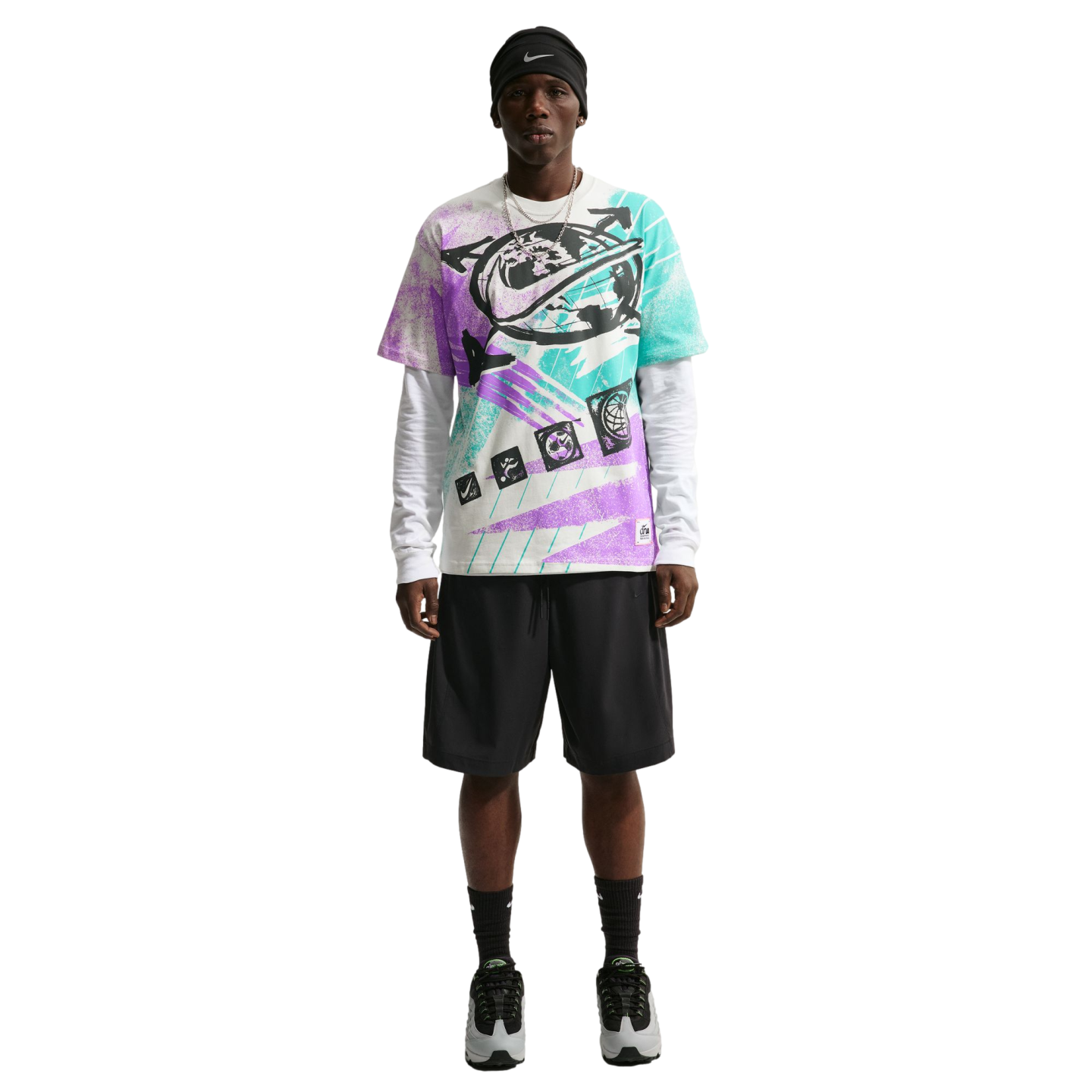 A person modeling a colorful purple, teal, and white graphic Nike t-shirt layered over a white long-sleeve shirt, paired with black athletic shorts, a black beanie, and grey sneakers against a solid black background.
