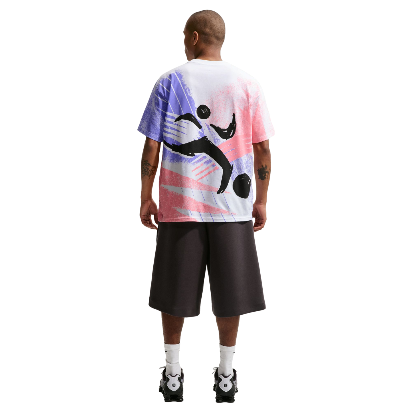 A rear view of a man standing against a black background. He is wearing a white oversized t-shirt with a large pink, purple, and black abstract graphic on the back, dark gray knee-length shorts, white crew socks, and black and white athletic shoes.