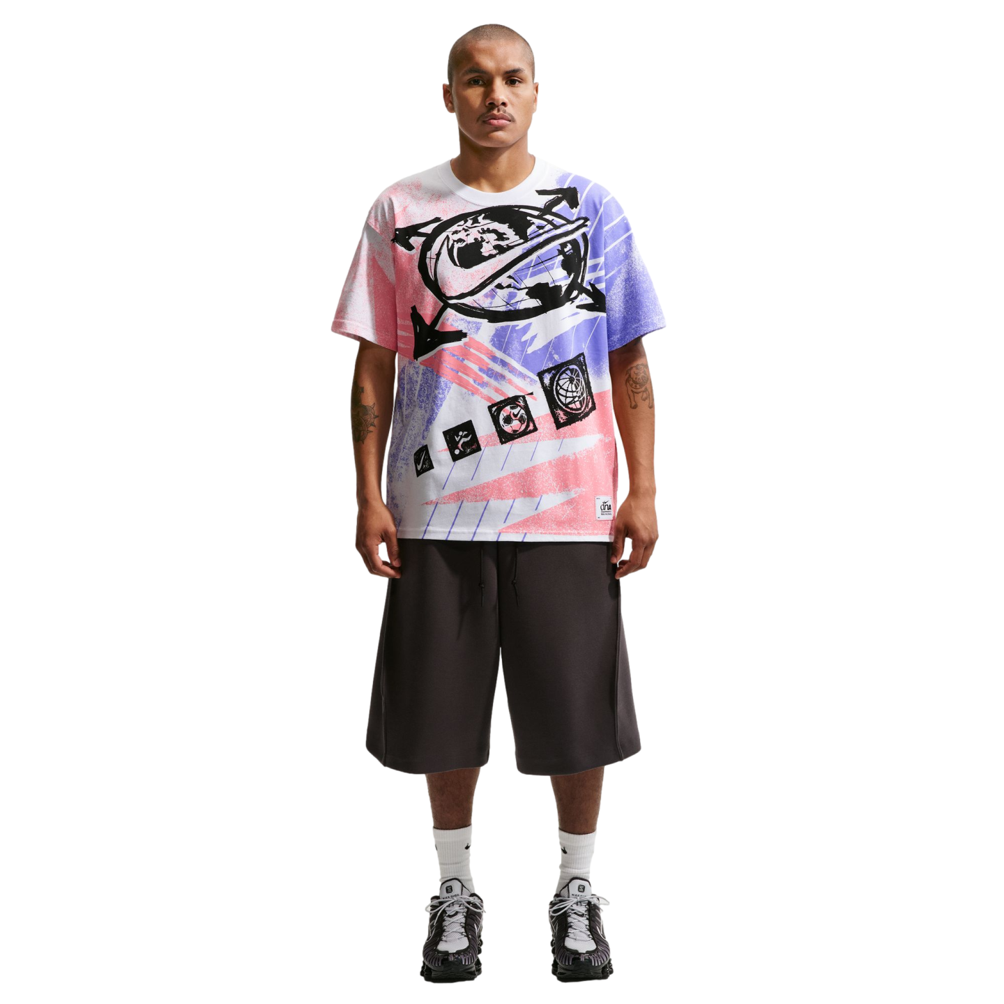 A full-length photo of a man standing against a black background, wearing an oversized graphic t-shirt with pink and purple spray-paint designs, wide-leg charcoal grey shorts, white crew socks, and grey athletic sneakers. He has short hair, a mustache, an
