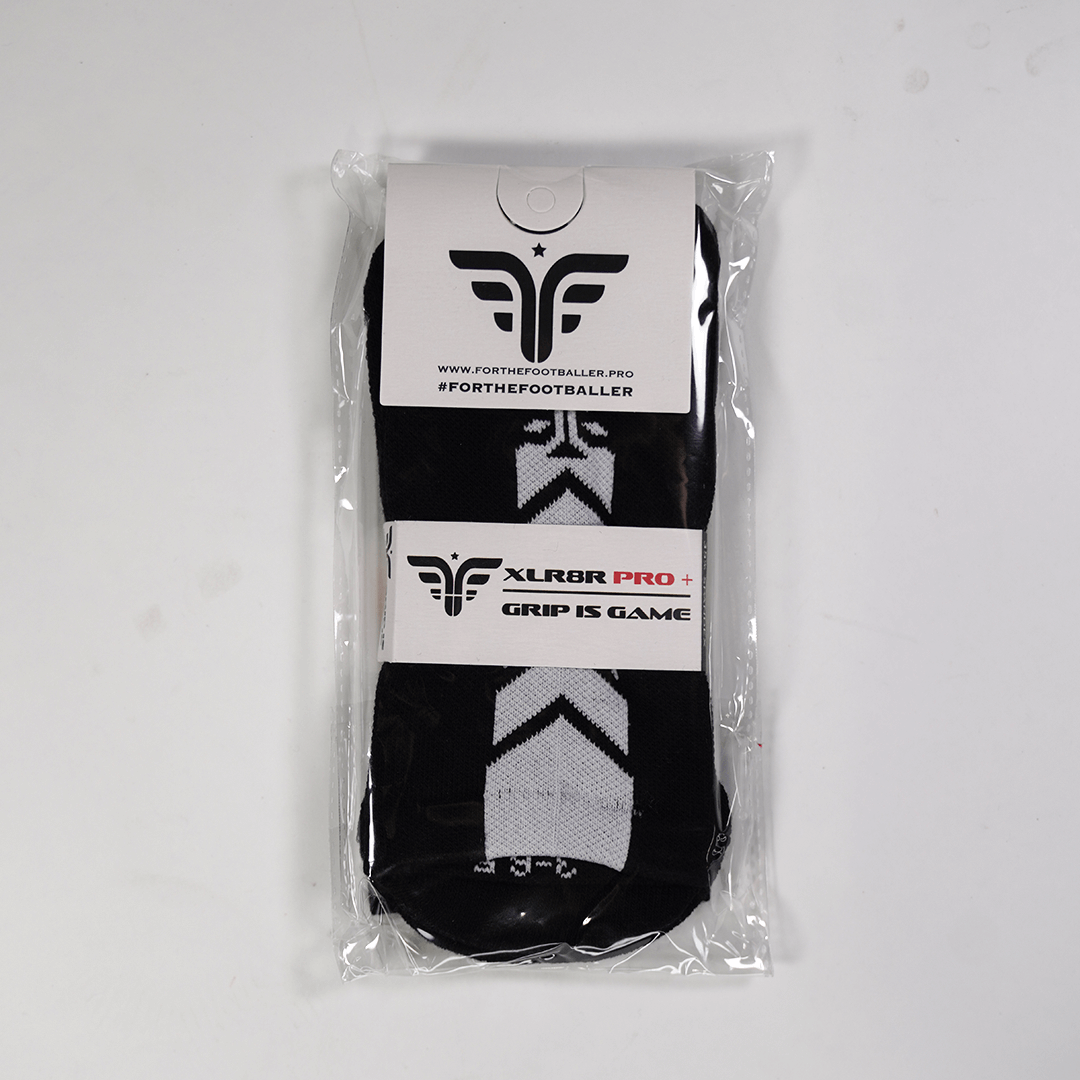 A pair of For The Footballer XLR8R Pro No Slip Socks - Black, shown in clear plastic packaging. The black socks feature white chevron-like patterns and are held by a white cardboard header and a middle band with the brand logo and "XLR8R PRO+" text.