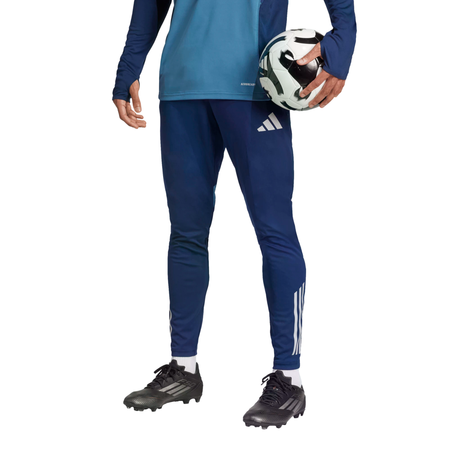 A person in blue athletic wear and black soccer cleats holds a soccer ball under their arm against a solid black background.